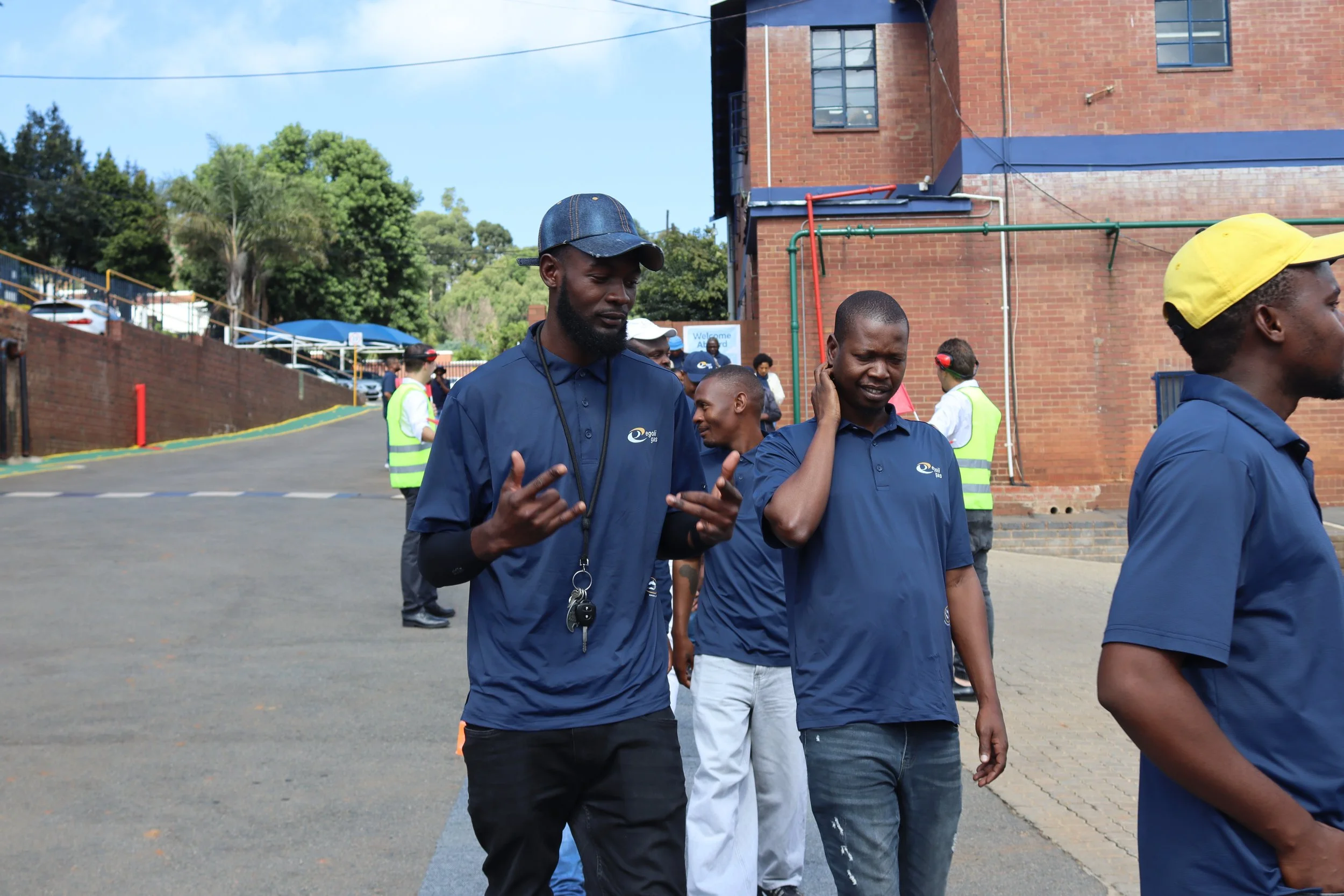 Group of men walking outdoors near a brick building with trees in the background, some wearing navy blue shirts with a logo, others in safety vests and hats.