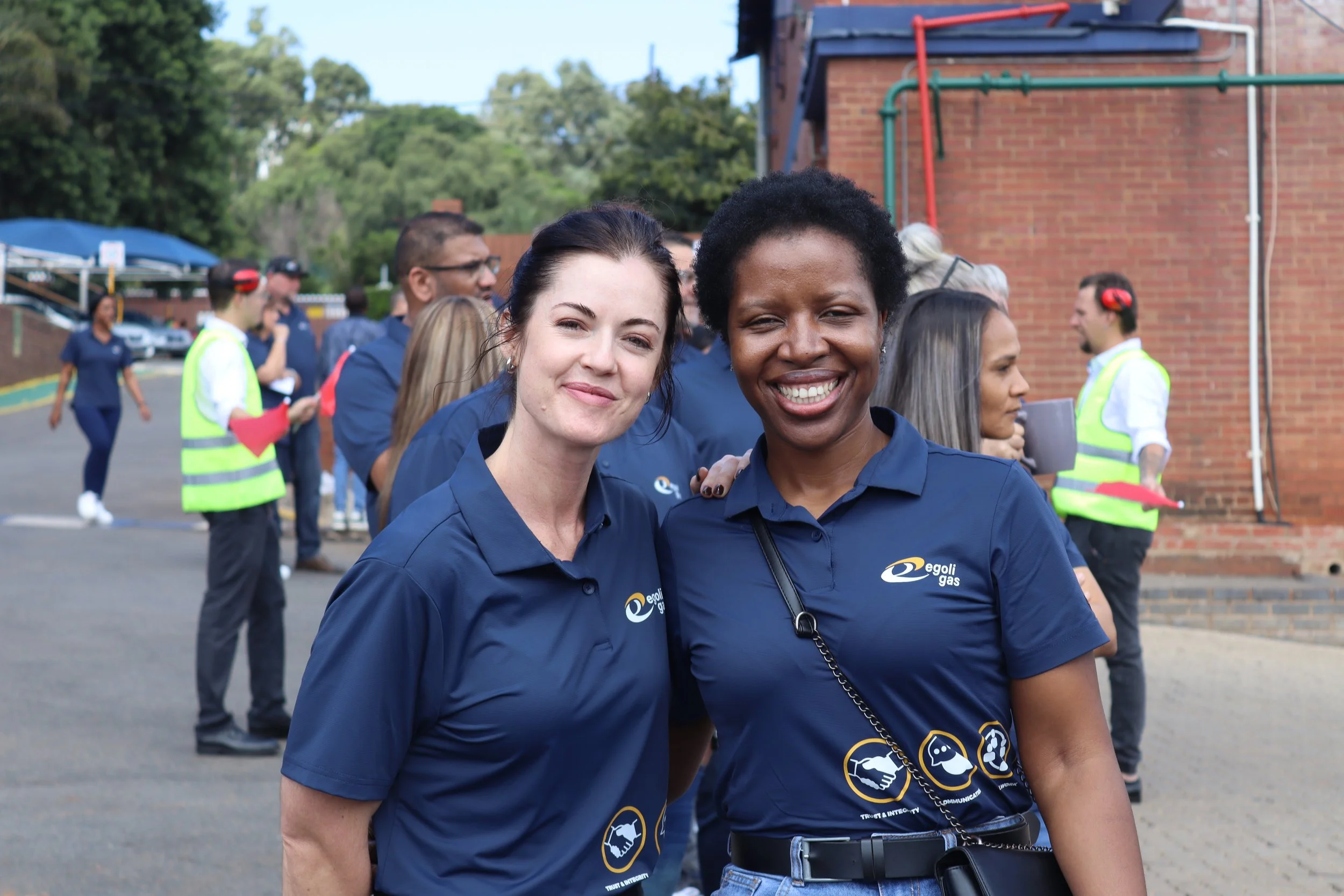 Two women smiling and standing together outdoors, wearing dark blue shirts with logos, with a group of people in the background.