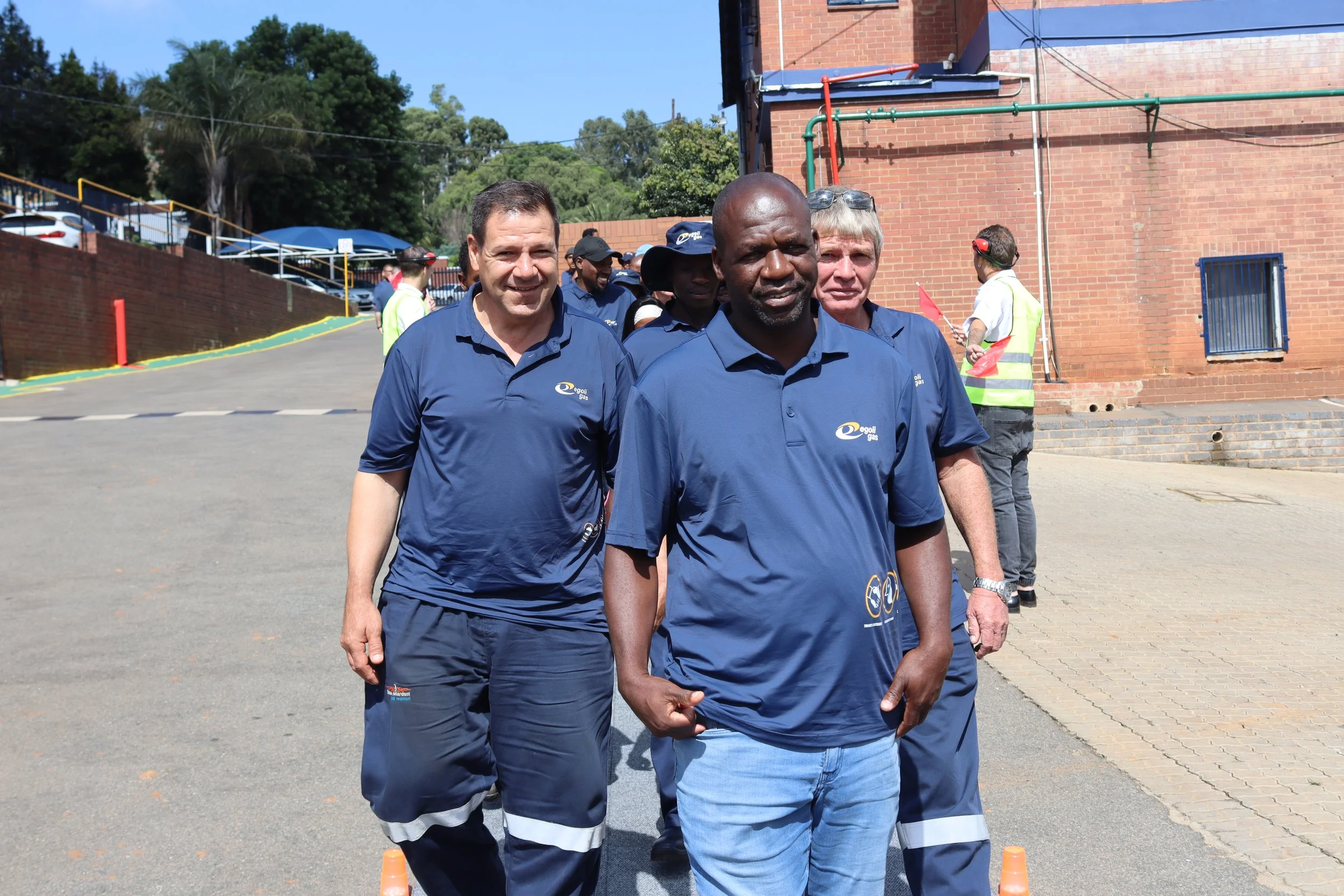 Three men in navy blue shirts walking outdoors, with other people and a brick building in the background.