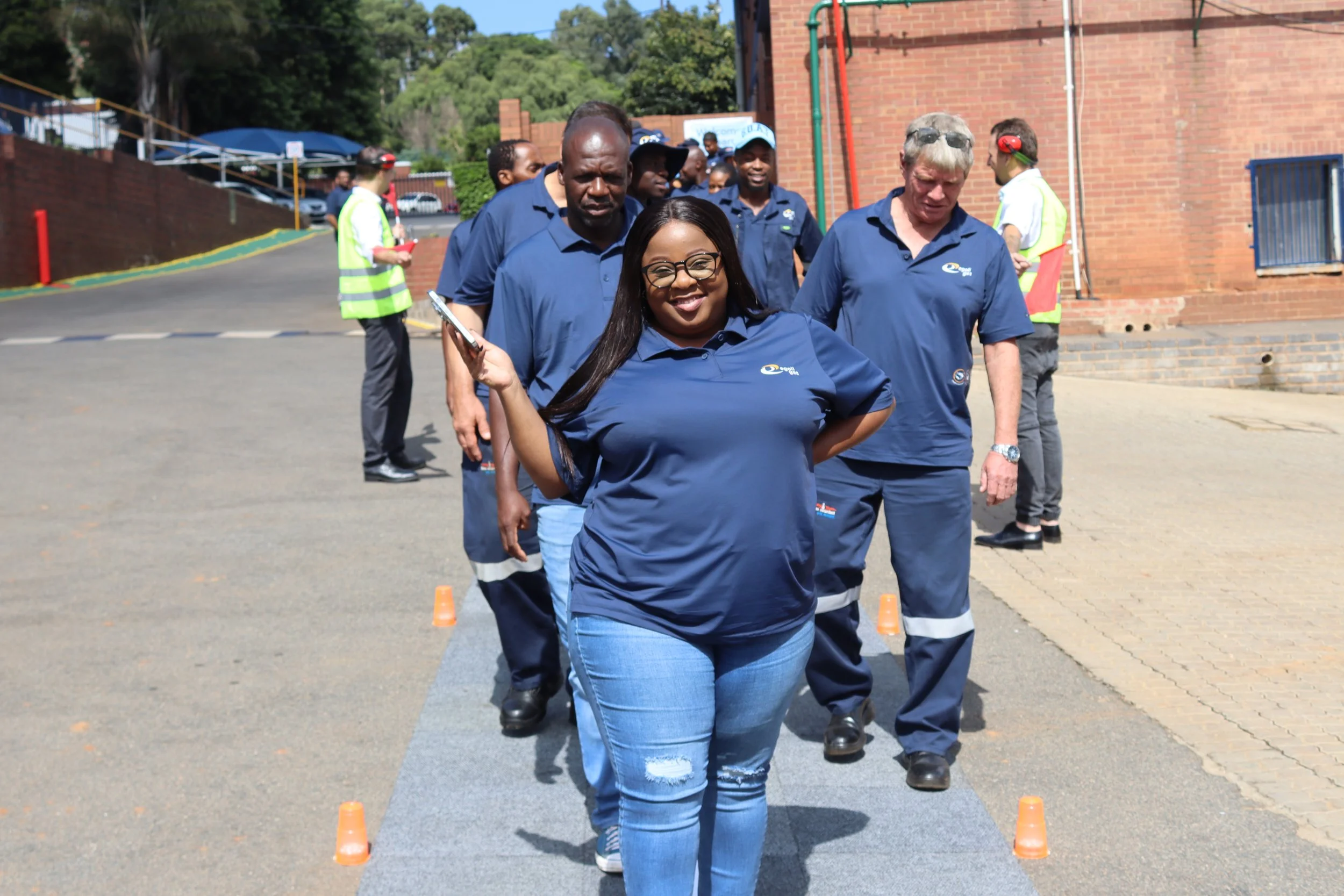 A group of diverse people participating in an outdoor team-building activity, walking behind a woman in blue jeans and a dark blue shirt, with orange traffic cones set up on the ground for a relay or obstacle course, in front of a brick building.