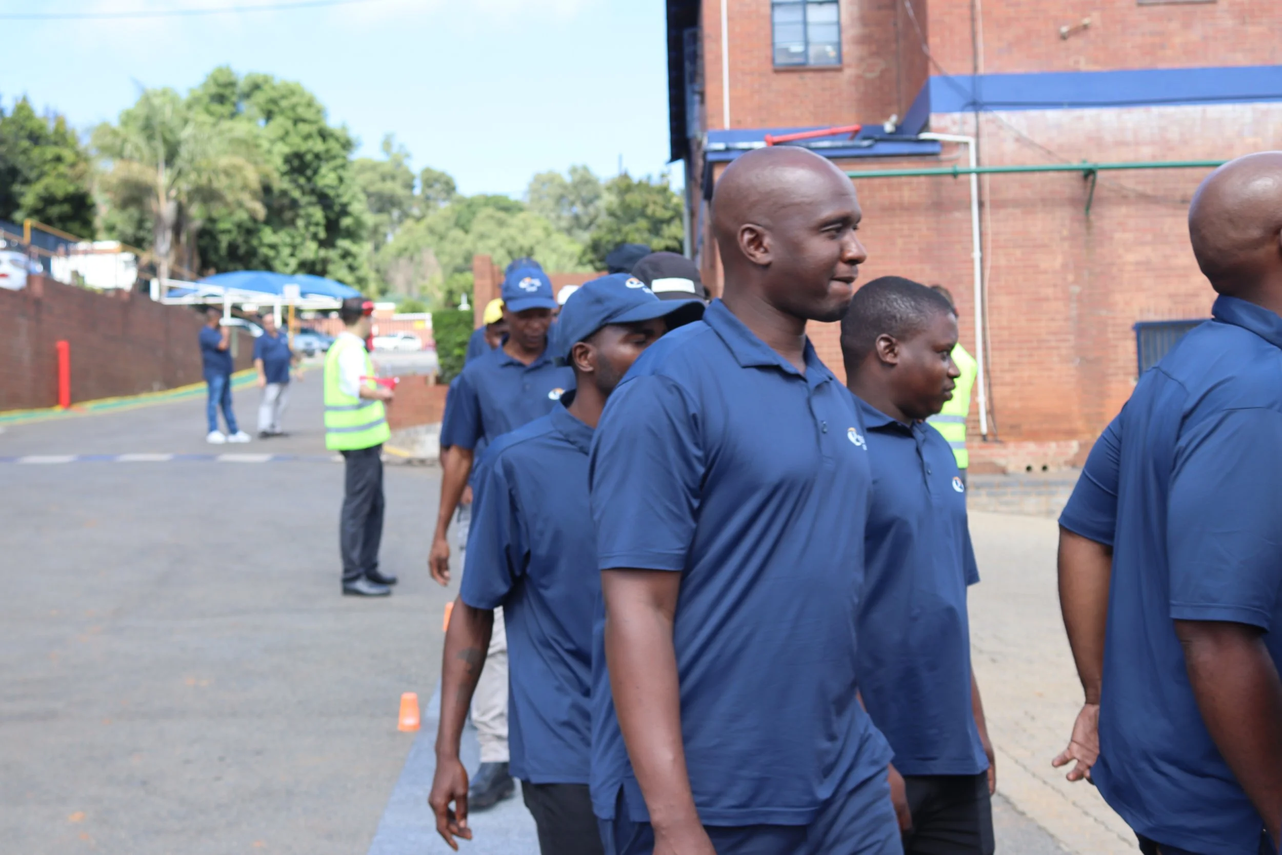 A group of men in navy blue shirts and caps walking in a line outdoors near a brick building with trees in the background.
