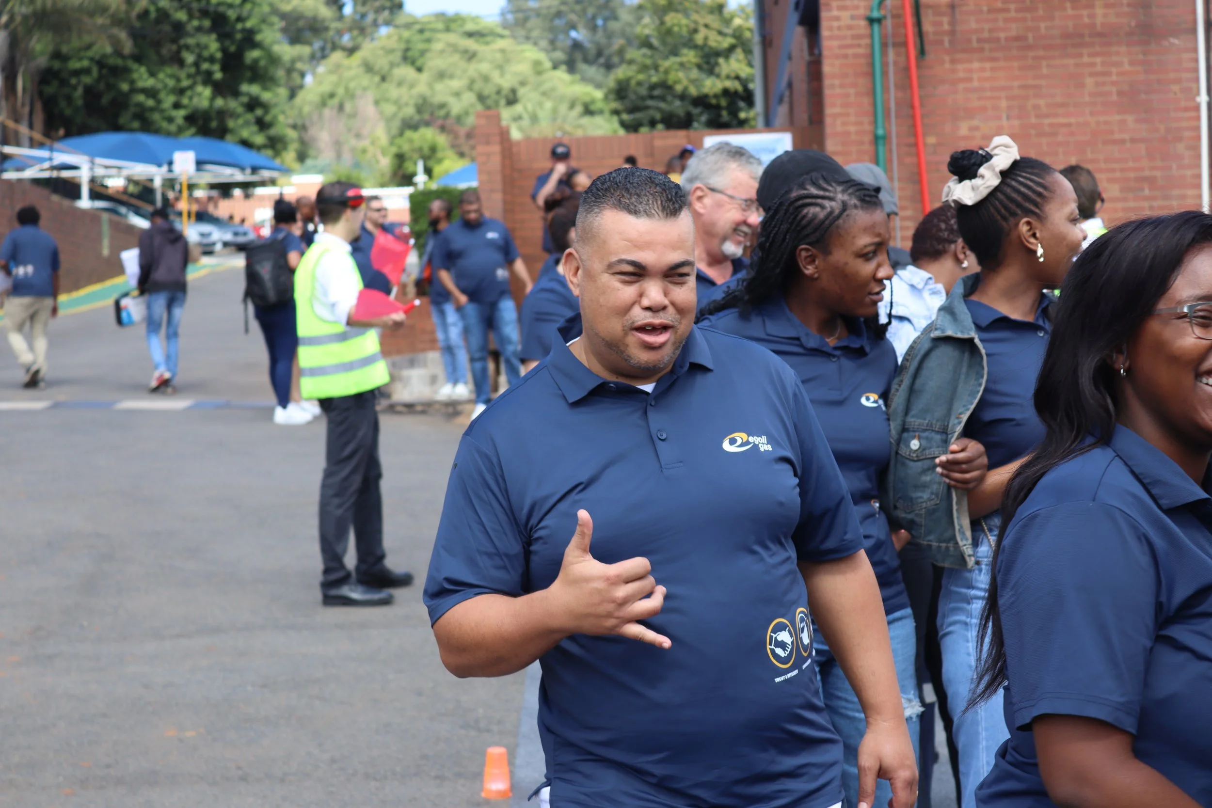 A group of diverse people wearing blue shirts and participating in an outdoor event, with some giving thumbs-up signs, standing behind traffic cones on a street with trees and brick buildings in the background.
