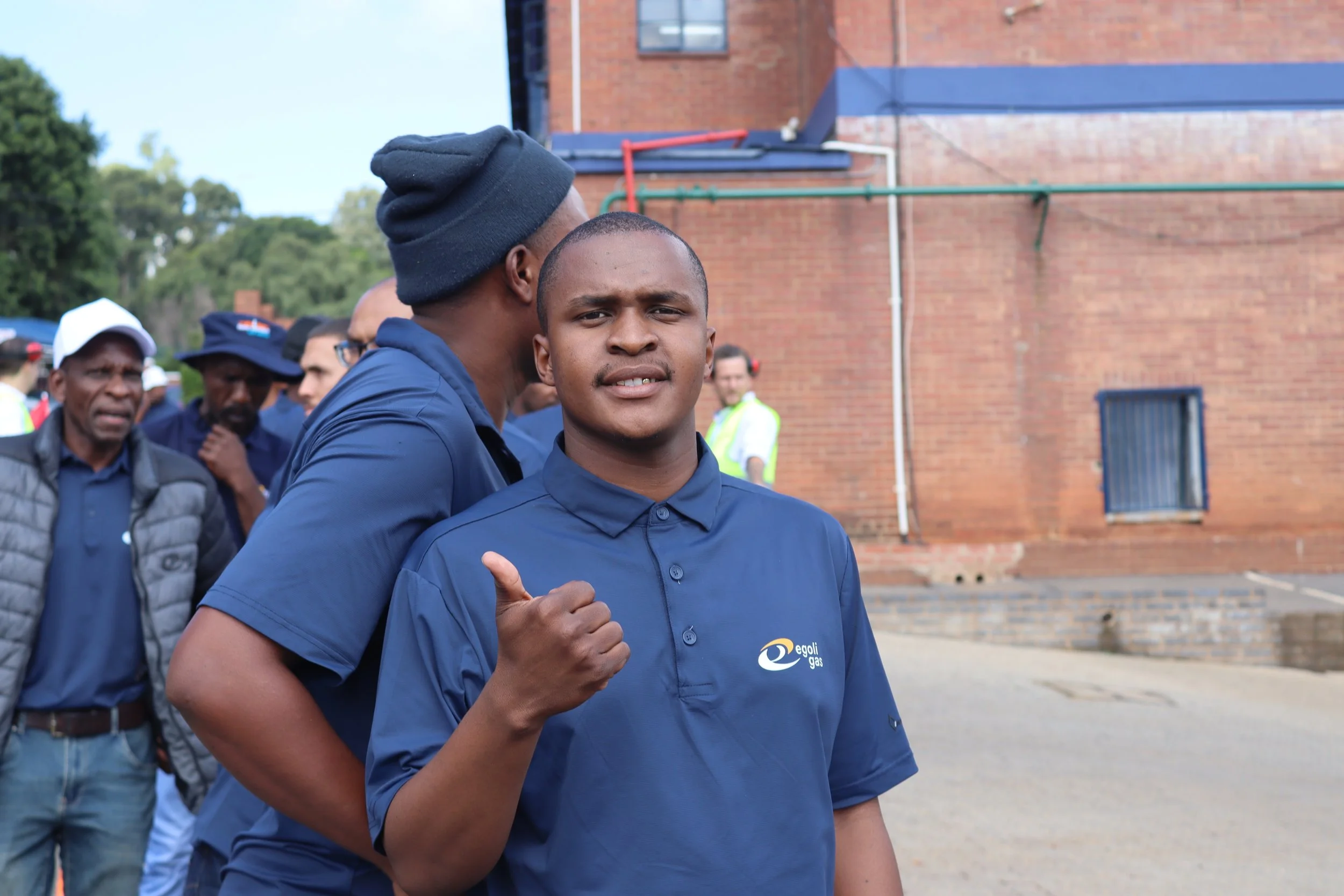 A young man in a blue collared shirt with a logo, giving a thumbs-up gesture, standing among a group of people outdoors near a brick building.