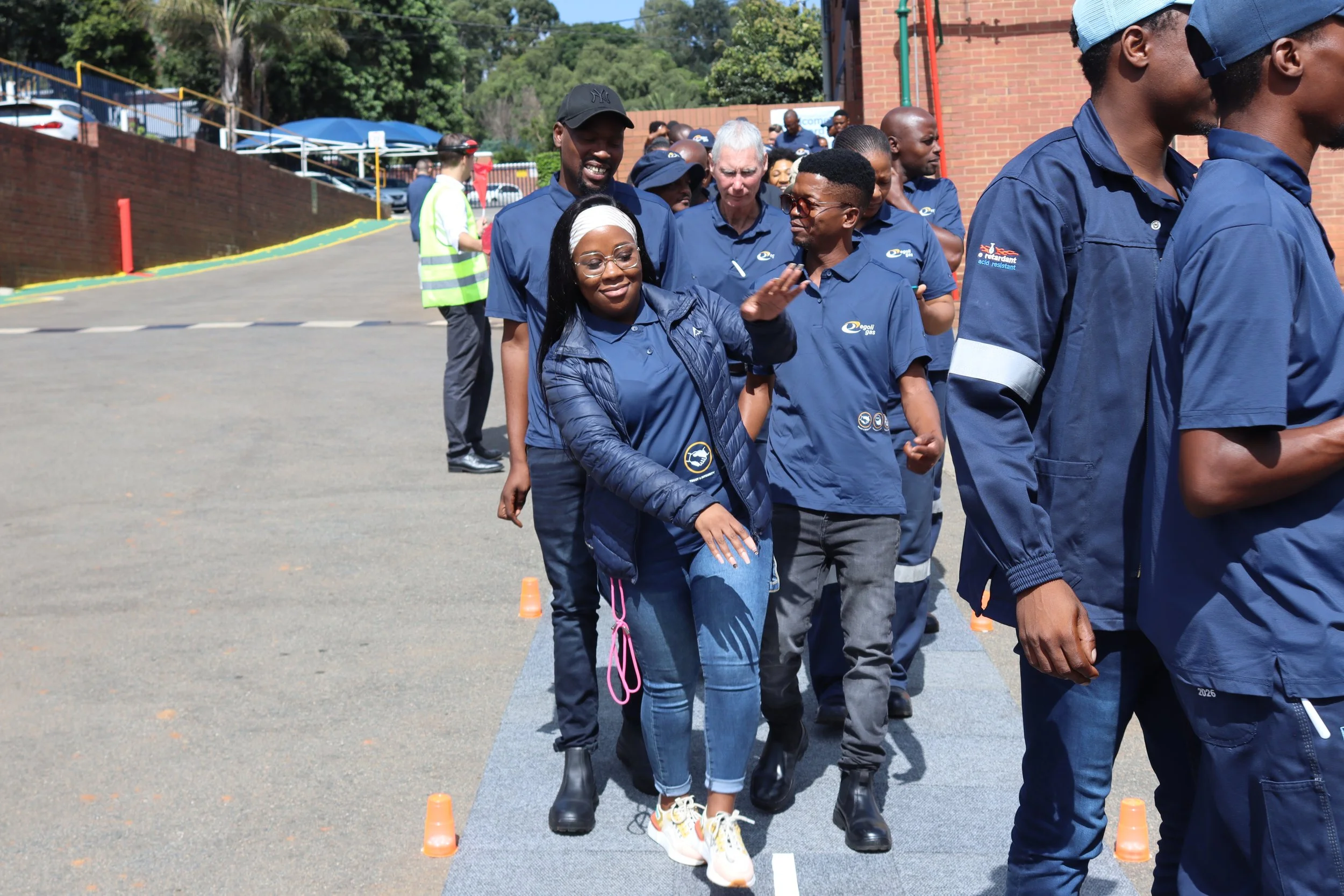 Group of people participating in an outdoor team-building activity, walking in a line between small orange cones on a paved surface, some wearing matching blue shirts.