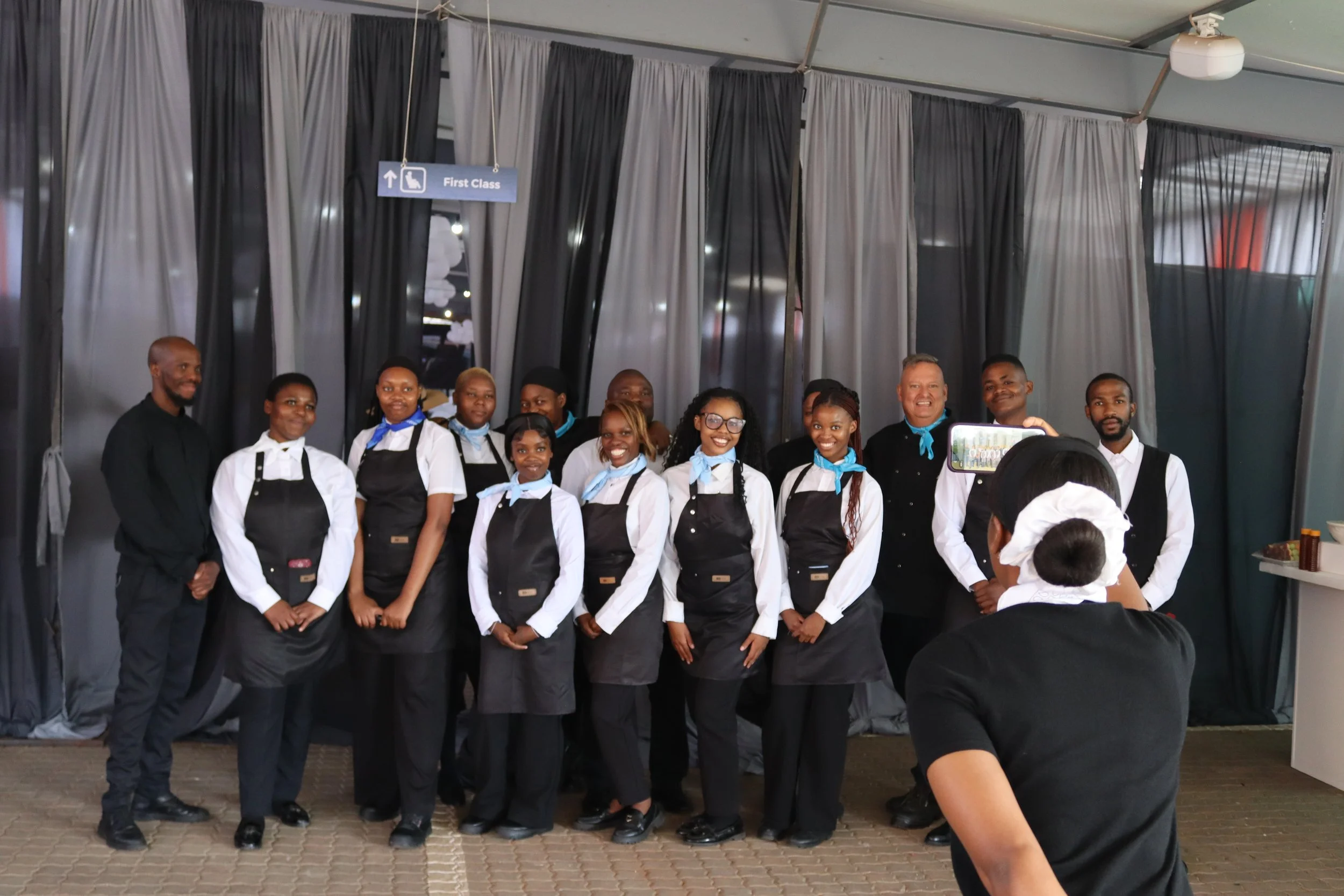 Group of eleven people, dressed in black and white uniforms with blue neck scarves, standing in front of a curtain at an airport. There is a sign above them that says 'First Class' with an arrow pointing upwards.