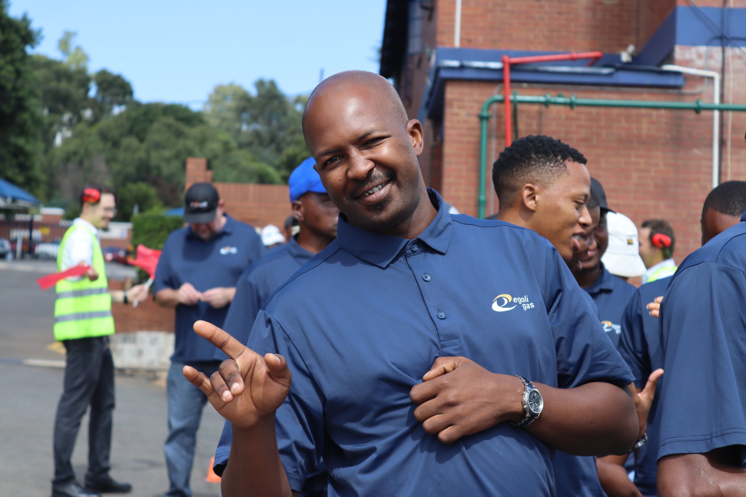 Group of people outdoors, some wearing blue shirts with 'egoli gas' logo, engaging in a group activity or dance, with trees and a brick building in the background.