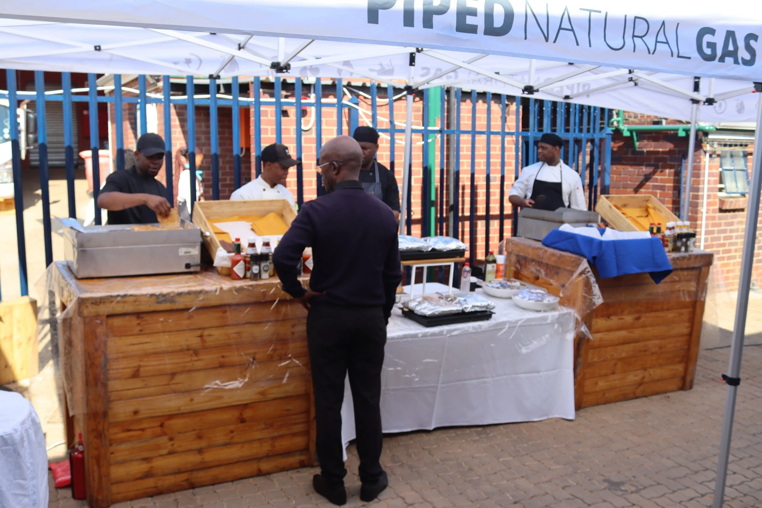A food stall at an outdoor event with a sign indicating natural gas-powered equipment. Several people, including two chefs in white and black uniforms, are working behind the wooden counter with bottles of condiments, and a customer is standing in fr