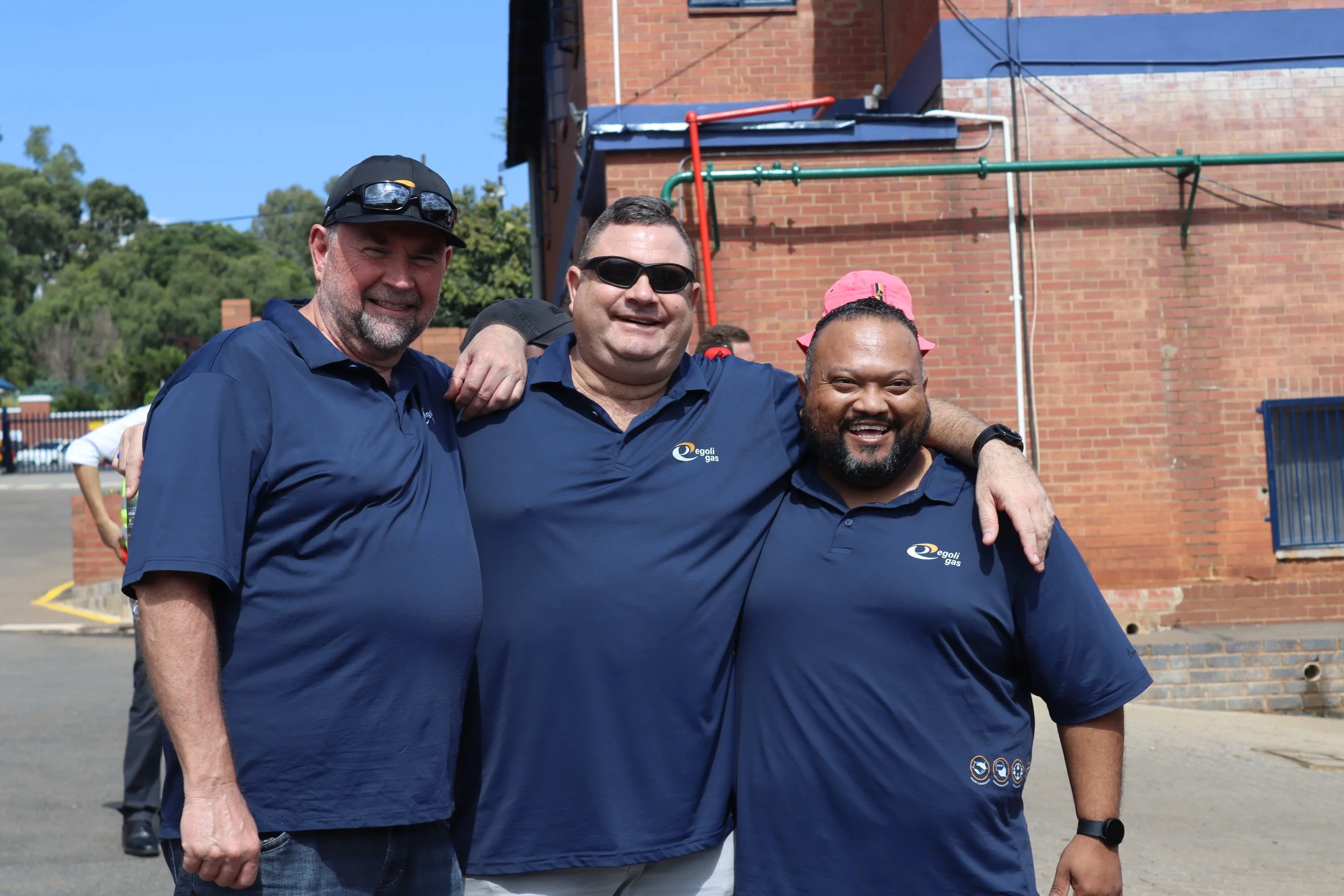 Three men smiling and standing close together outdoors, with one man’s arm around another's shoulders, in front of a brick building. They are wearing dark blue shirts with a logo reading 'egoli gas'.
