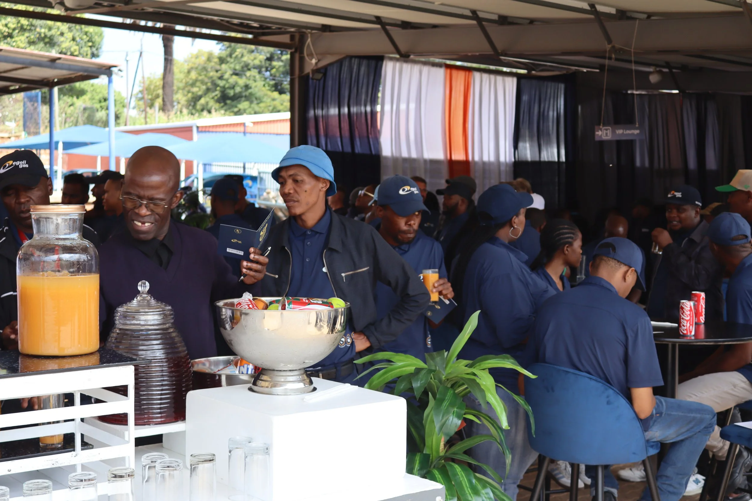 A group of people gathered at an outdoor event, some wearing blue uniforms and caps, with a buffet table with drinks and food in the foreground, and a backdrop of trees and a building with orange and blue curtains.