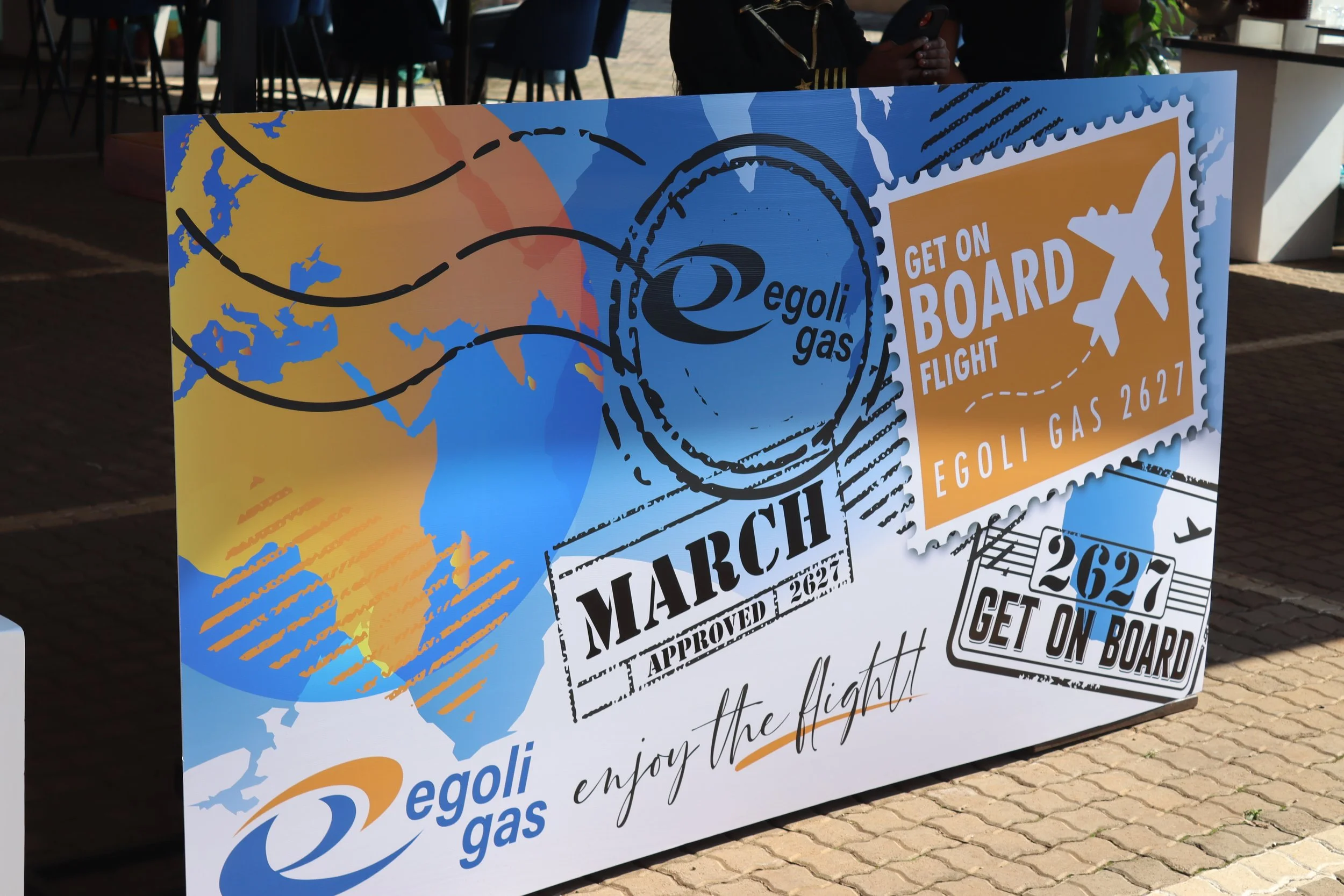 Large colorful sign promoting Egoli Gas with travel-themed graphics including stamps, airplane icons, and a world map, located outdoors on a brick-paved area.