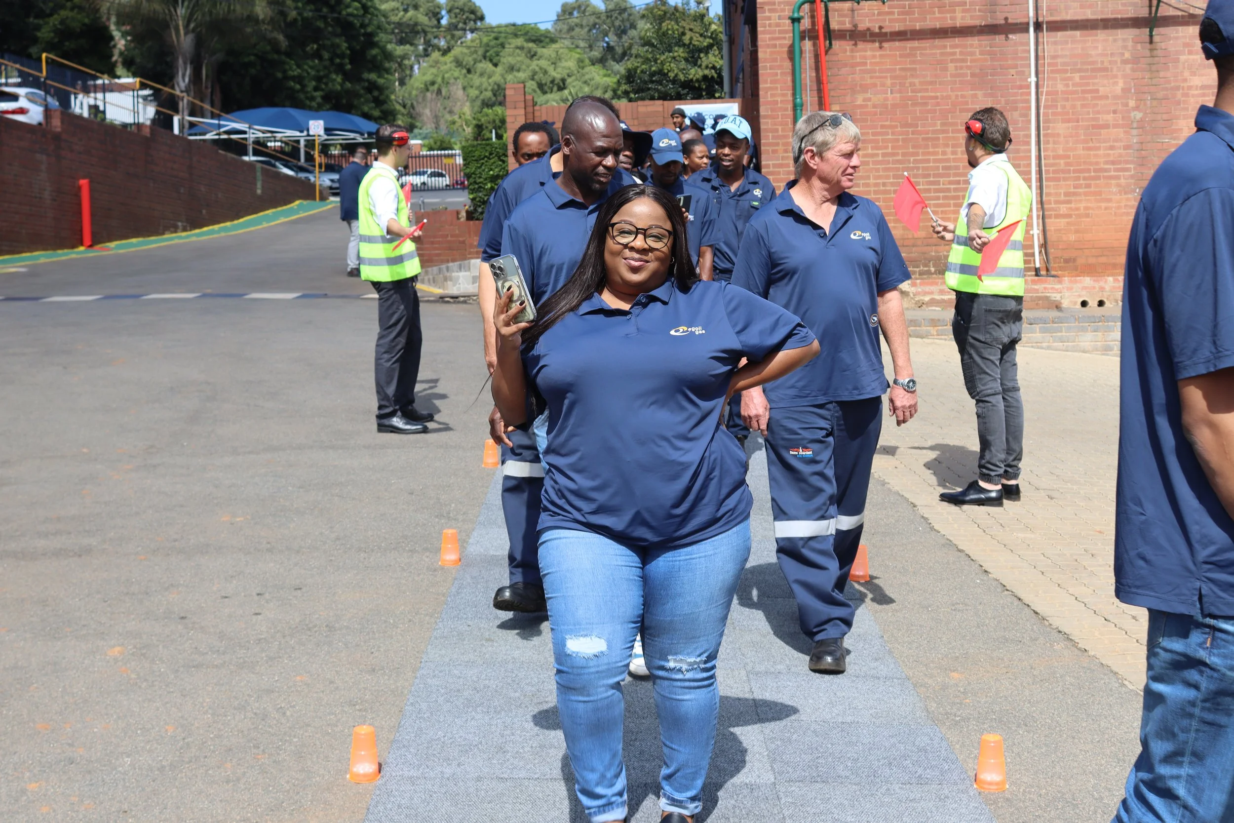 Group of people participating in an outdoor event, wearing blue uniforms, with some holding or near orange cones, on a paved area next to a brick building.
