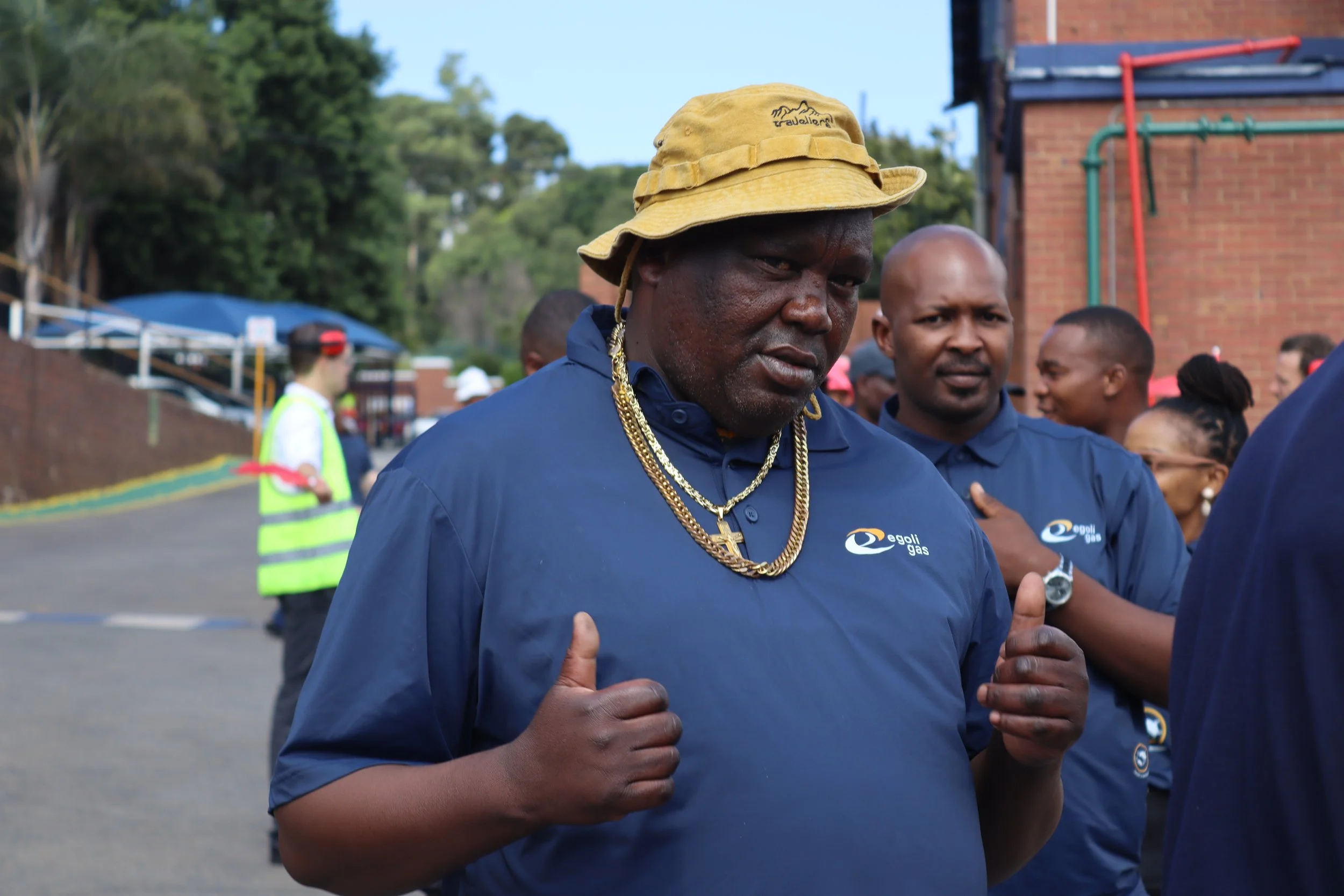 A man with dark skin wearing a yellow bucket hat, gold chains, and a navy blue shirt with a logo, standing outdoors among a group of people, some of whom are also wearing navy blue shirts. The background shows trees, a brick building, and other peopl