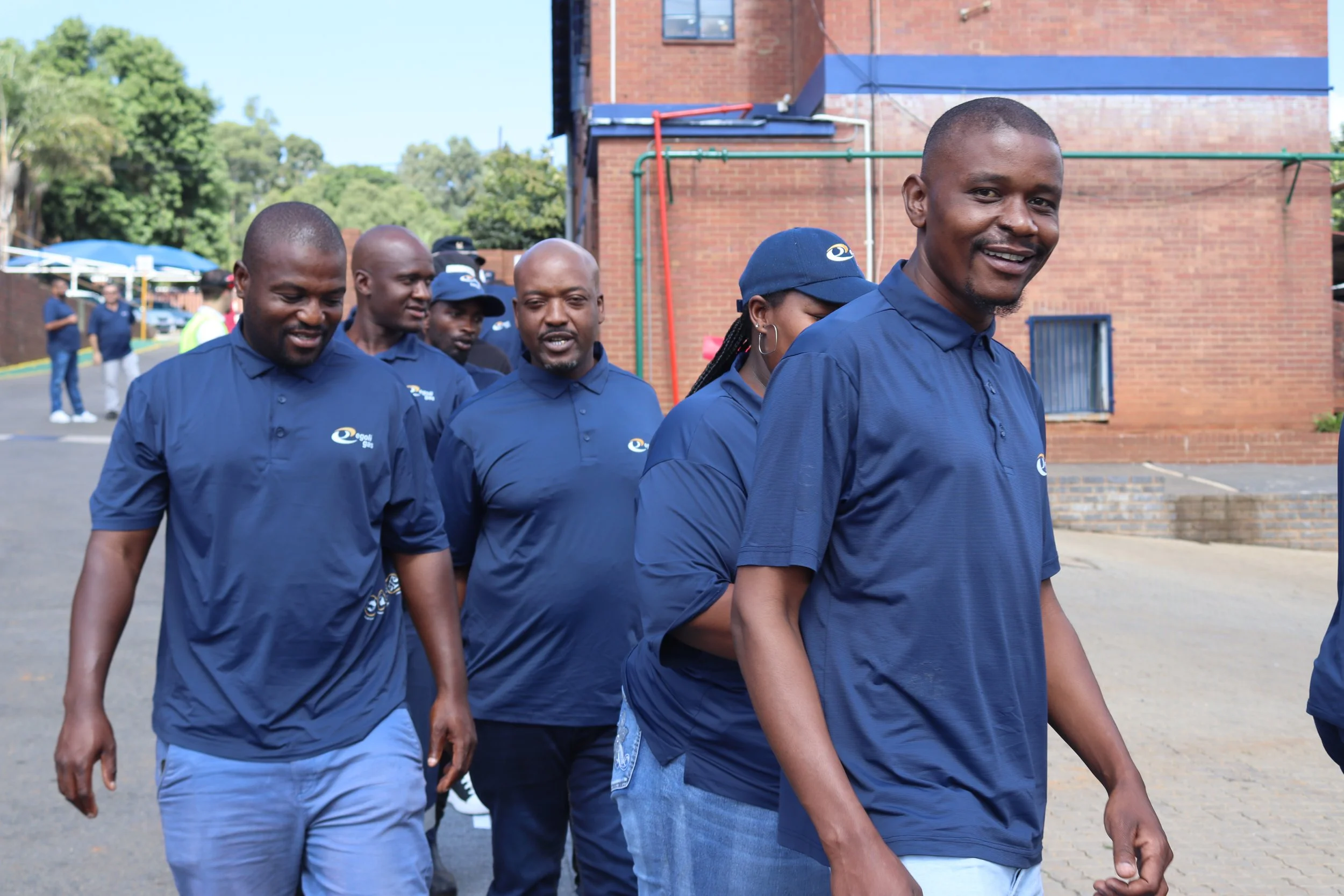 A group of people walking outdoors near a brick building, wearing navy blue shirts with a logo, some with hats or caps, and smiling.