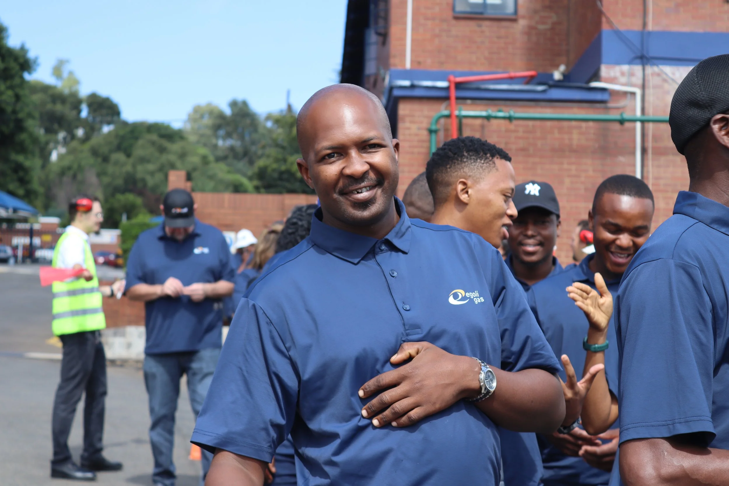 Group of people, mostly men, wearing blue shirts with 'Egoli Gas' logo, engaging in an outdoor activity on a sunny day. Some are smiling, talking, and shaking hands, with a brick building and trees in the background.