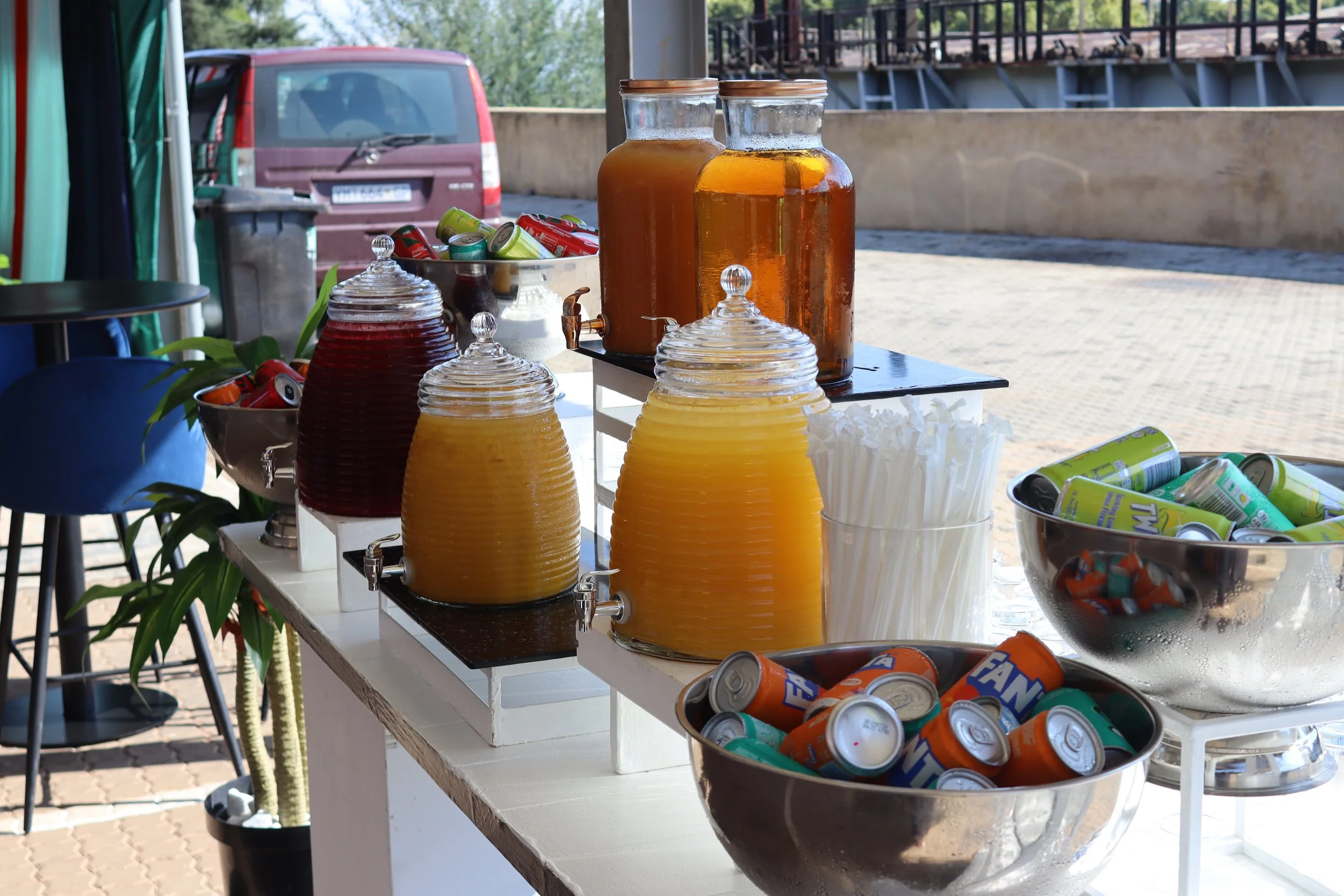 Table with various beverages including orange juice and iced tea in jars, and cans of soda, set outdoors with a parking lot and metal fence in the background.