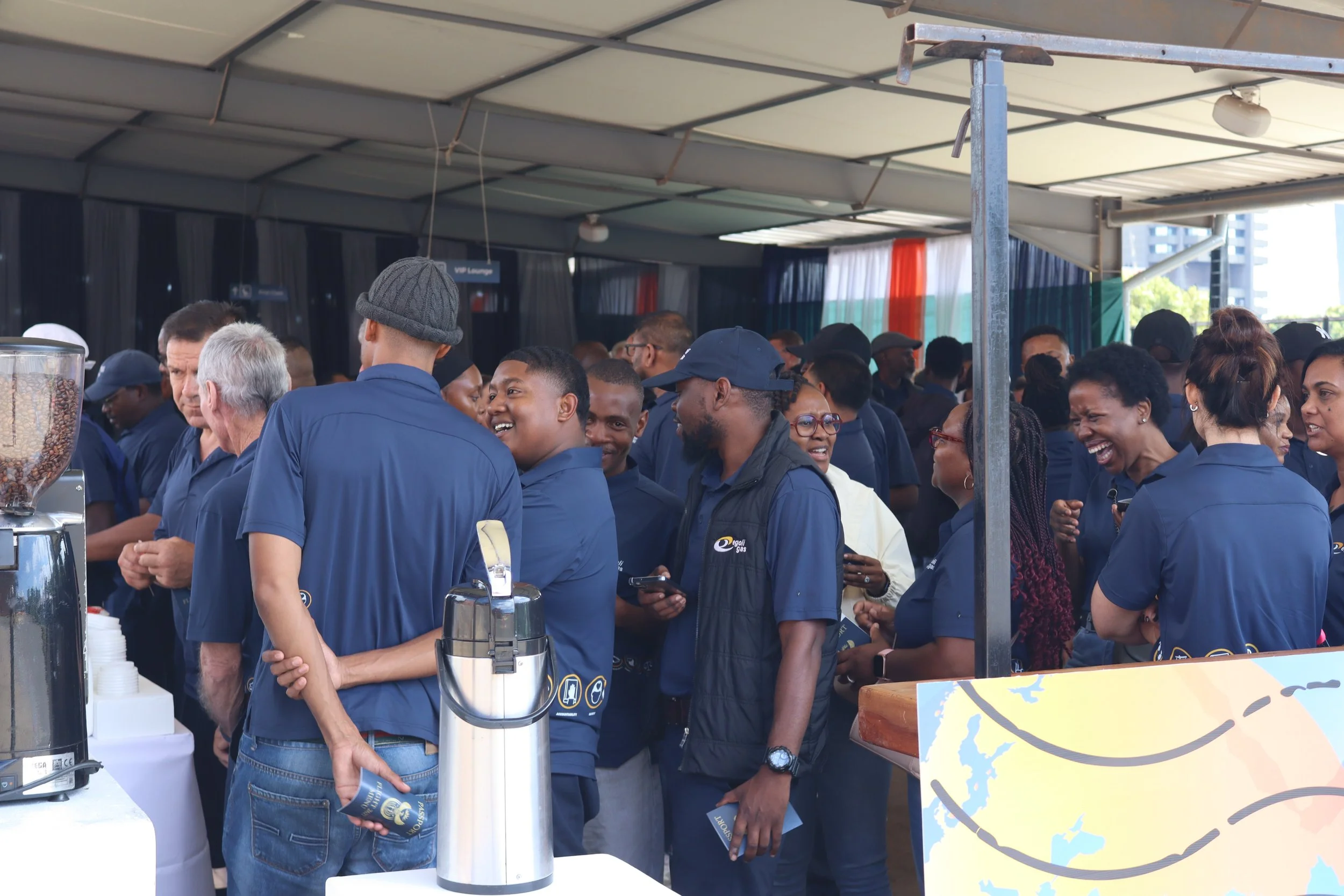 Crowd of people gathered at an outdoor event, some laughing and chatting, under a large canopy, with visible coffee dispensers and a colorful map in the foreground.