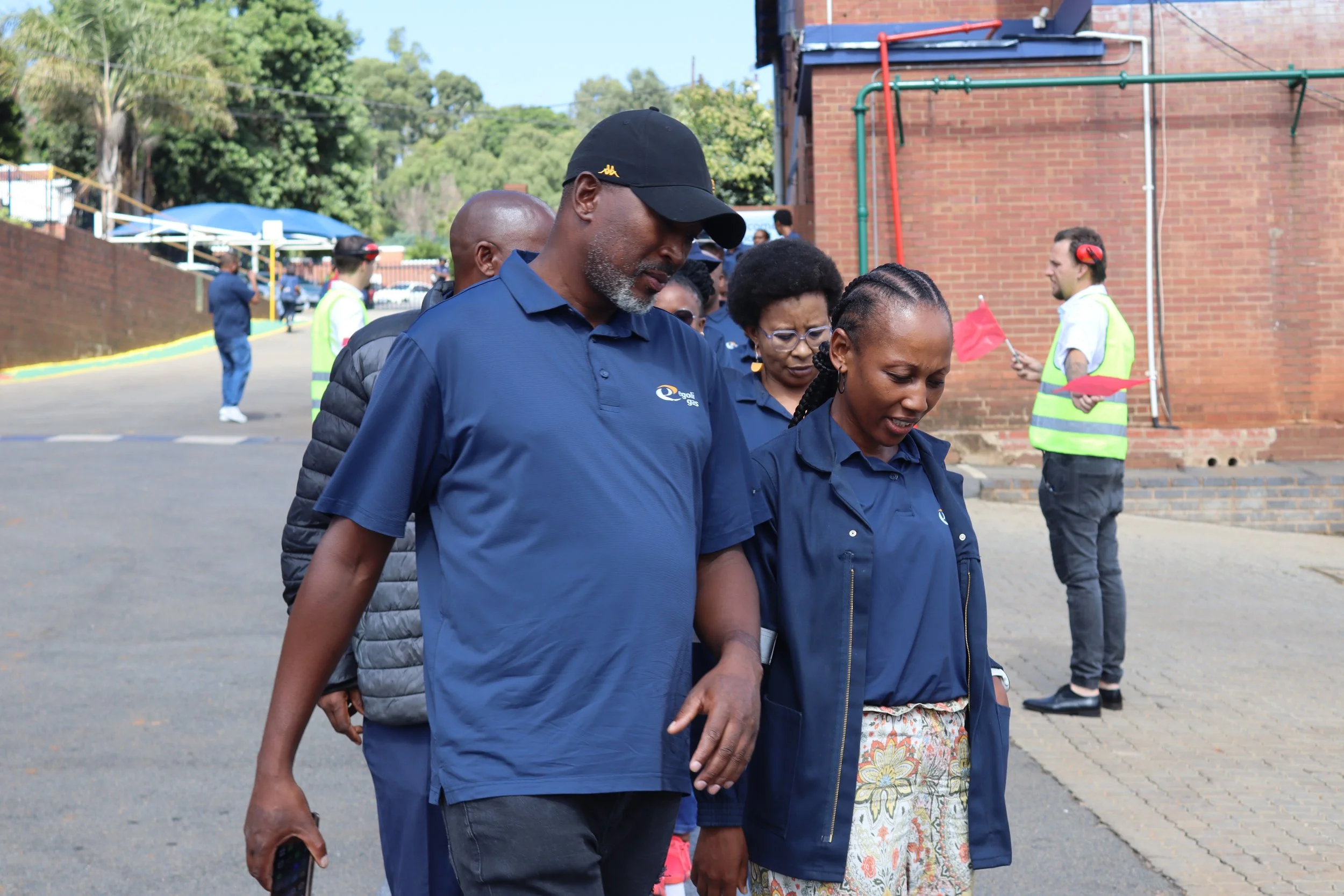 Group of people walking outdoors near a brick building, with some wearing blue uniforms, one holding a phone.