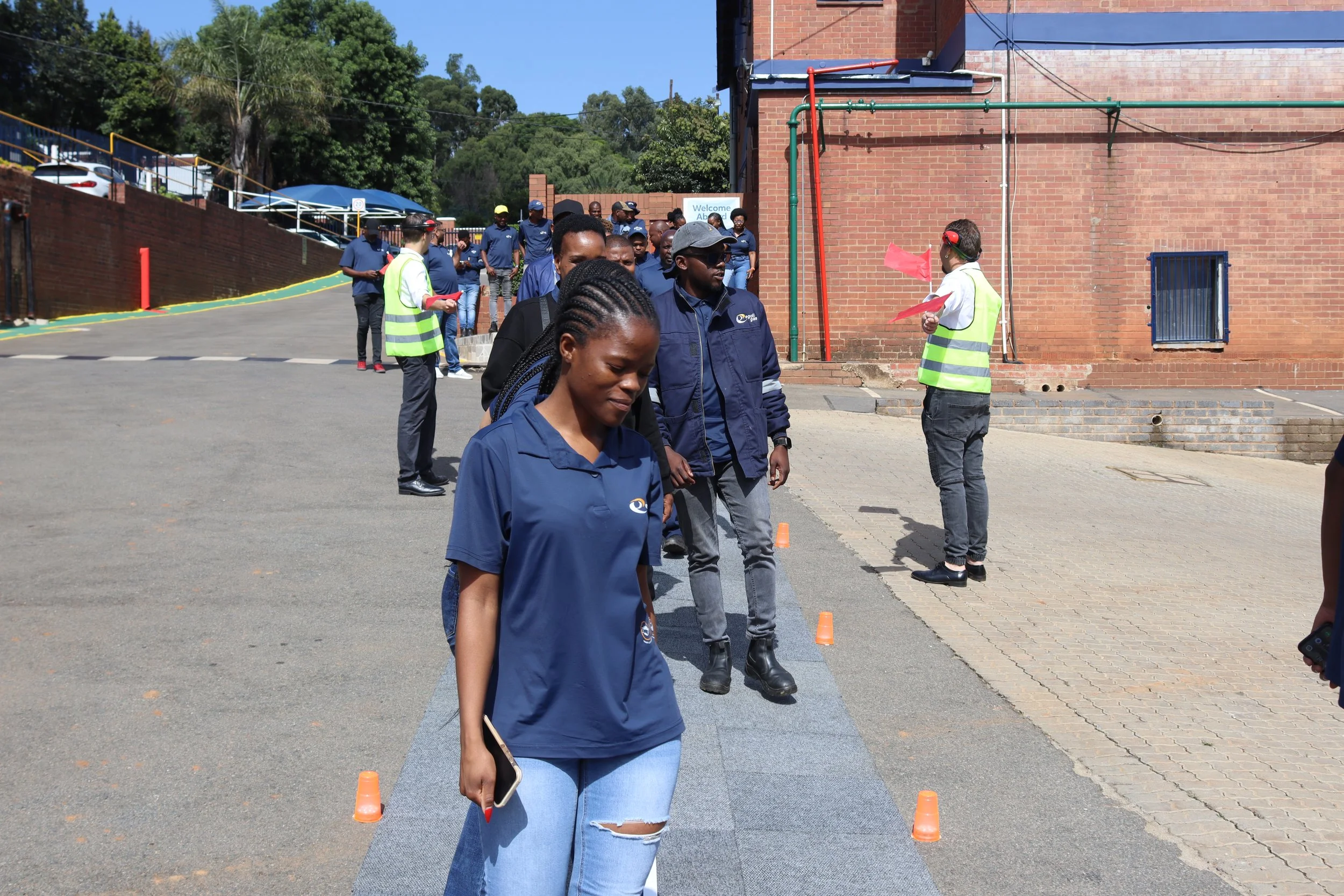 A diverse group of people participating in an outdoor event, some wearing safety vests, standing on a gray mat lined with orange cones, next to a brick building with pipes and a blue sign that says "Welcome Abed." There are trees and parked cars in t
