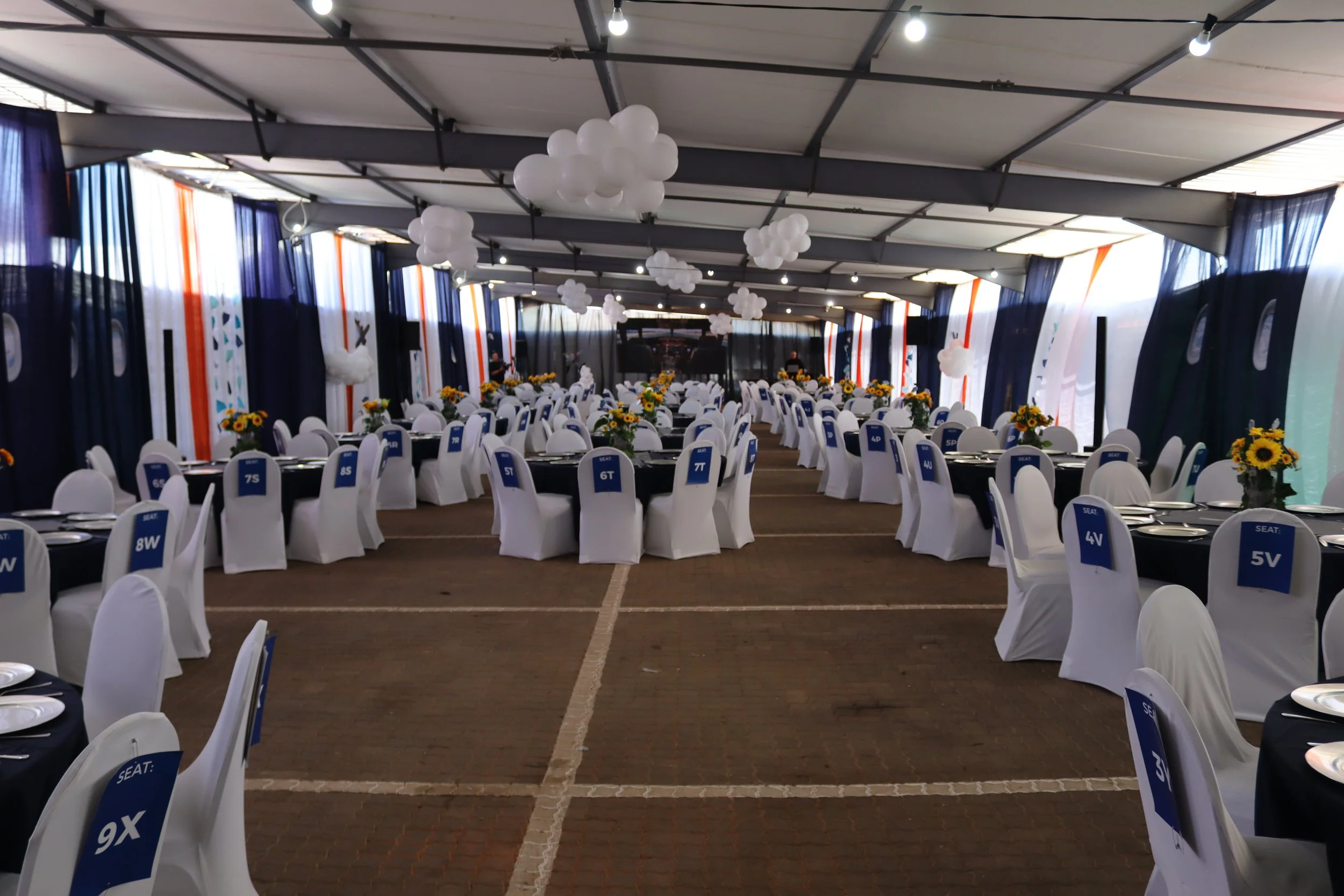 A large event tent decorated with white balloons hanging from the ceiling, navy blue and orange curtains along the sides, and round tables covered with white chair covers and dark tablecloths, each with seat number signs and sunflower centerpieces.