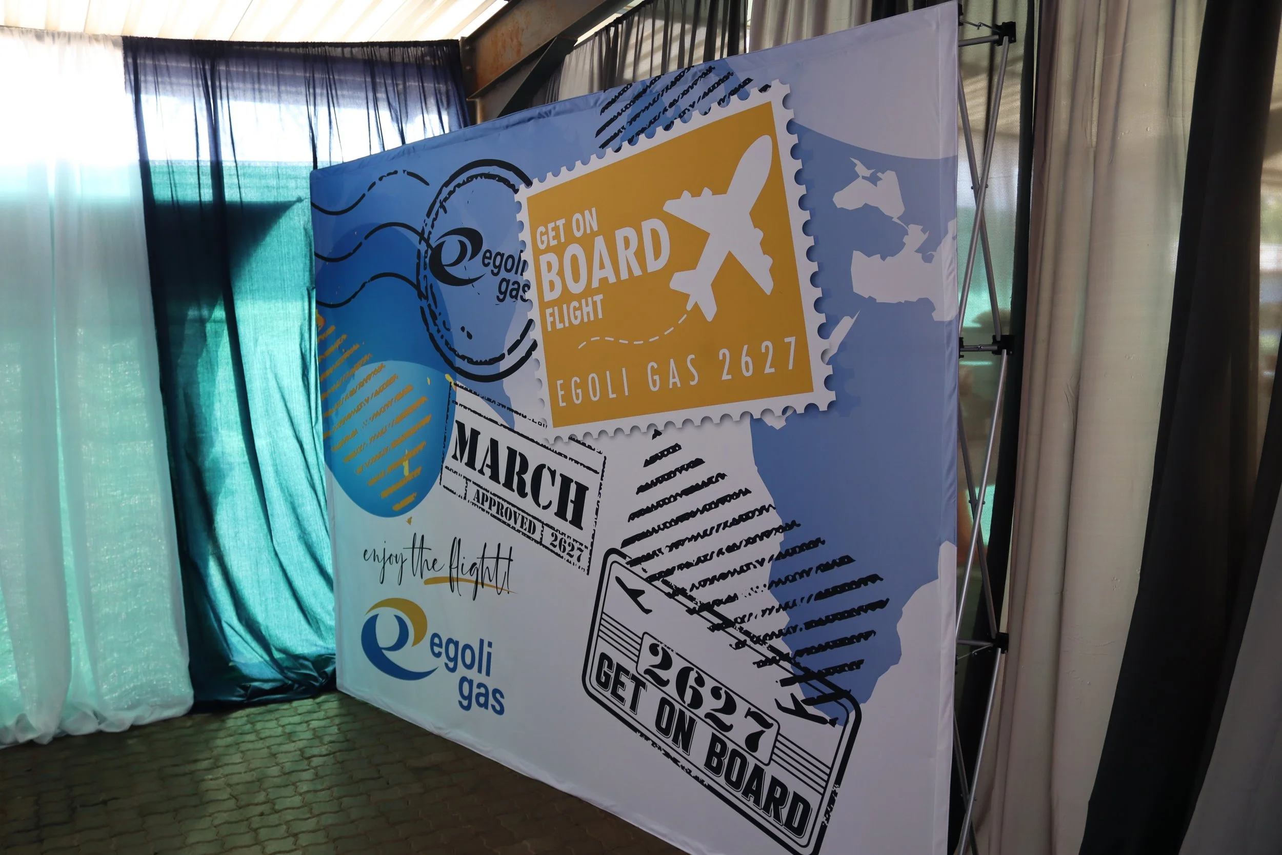 Poster promoting Egoli Gas with a graphic of an airplane, stamp-like graphics, and various travel-themed illustrations, with curtains and a window in the background.
