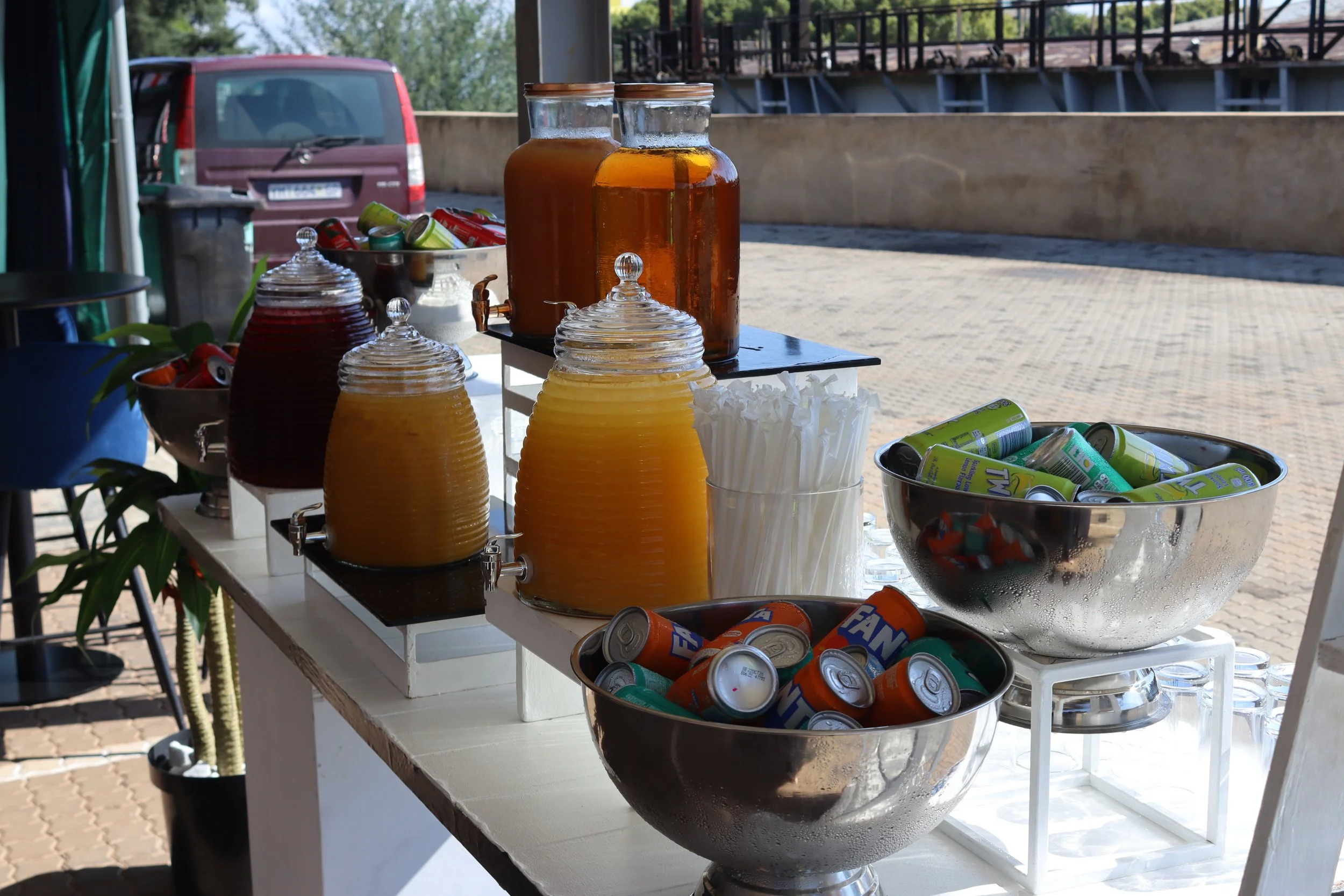 Table with large glass containers of orange and brown beverages, glasses, canned drinks, and snacks in containers, set outdoors with a parking lot and trees in the background.