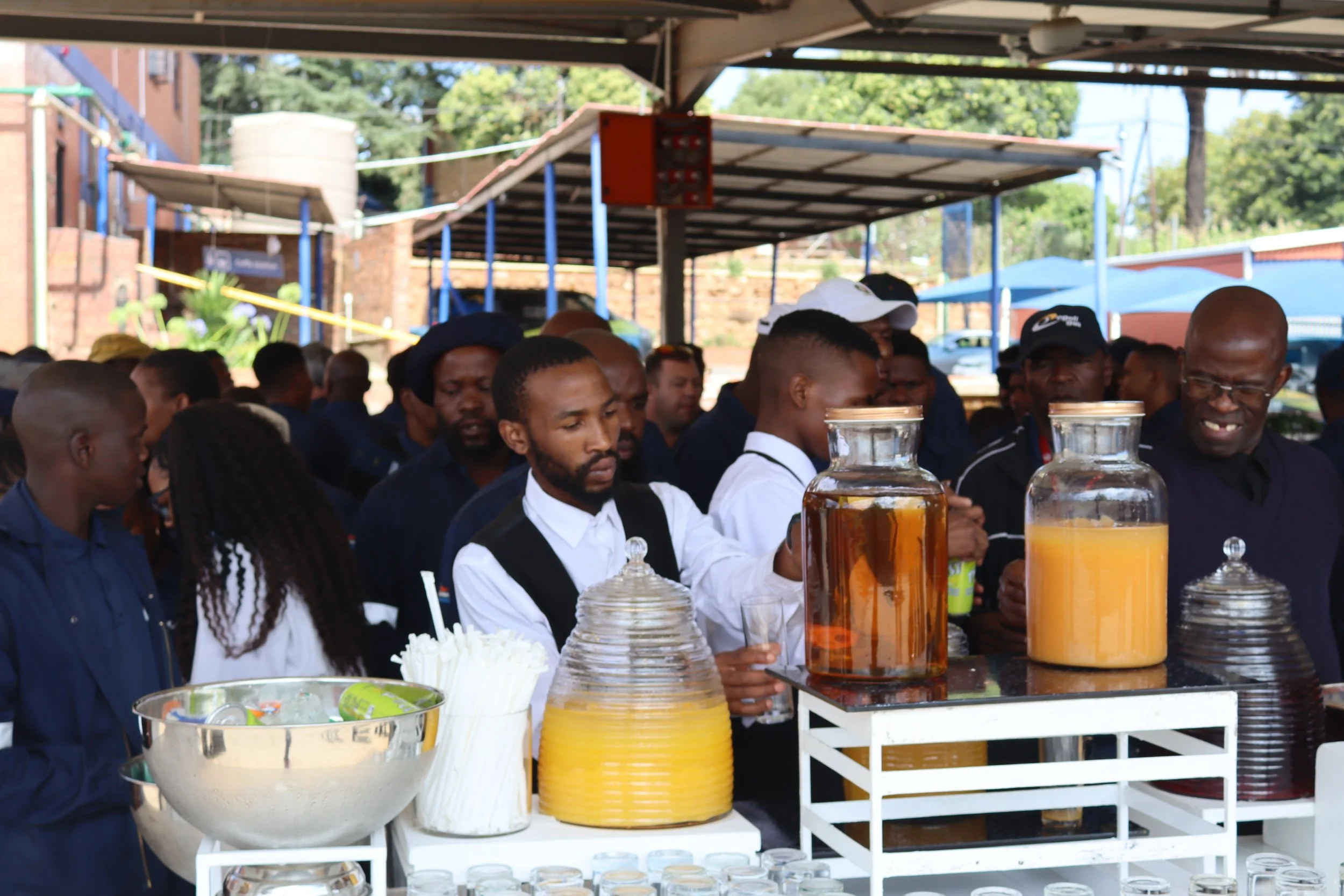People gathered at a drink stand outdoors, with large jars of beverages and cups on display.
