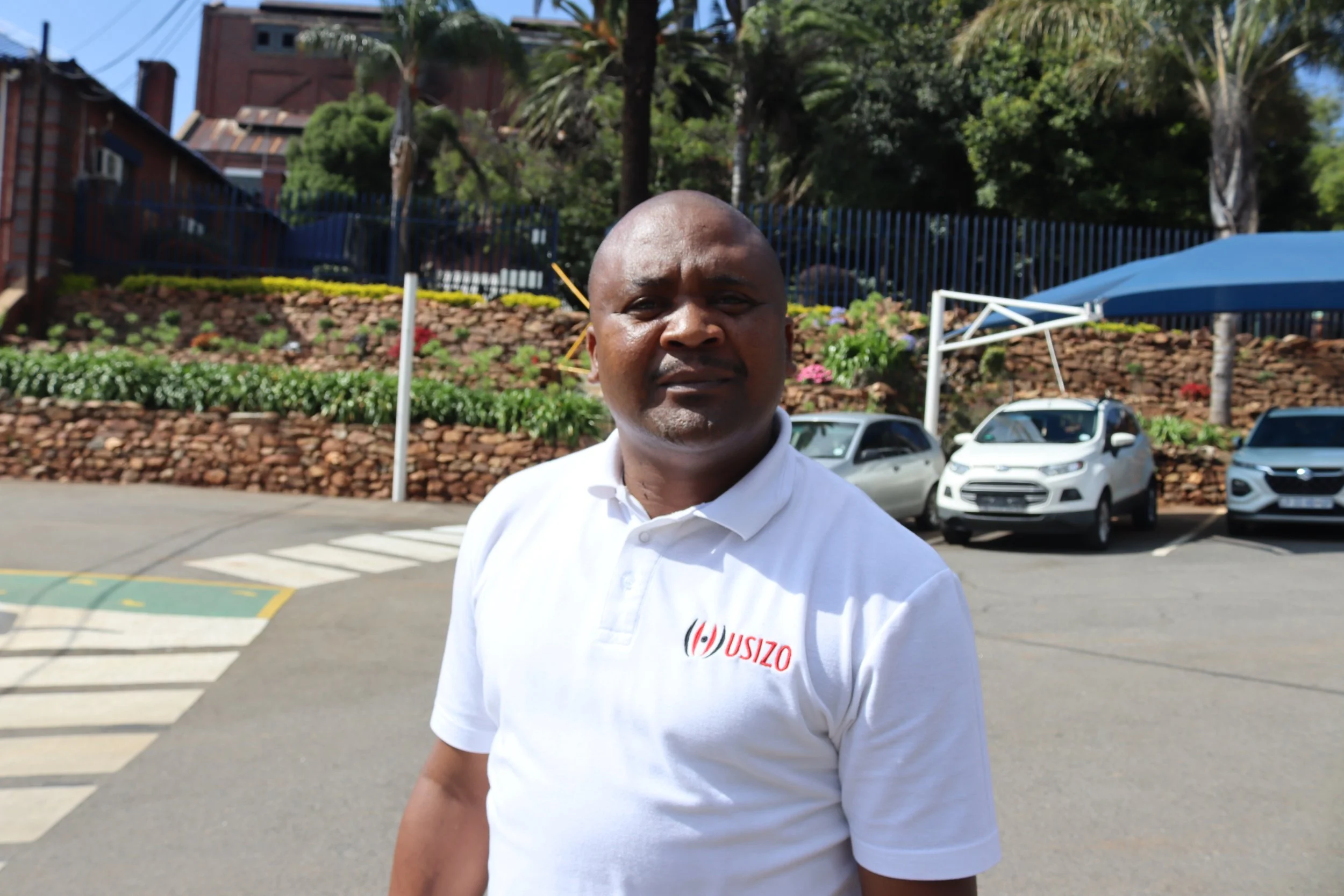 A man in a white polo shirt standing outdoors in a parking lot, with a beautiful garden with flowers and trees behind him, and several parked cars nearby.
