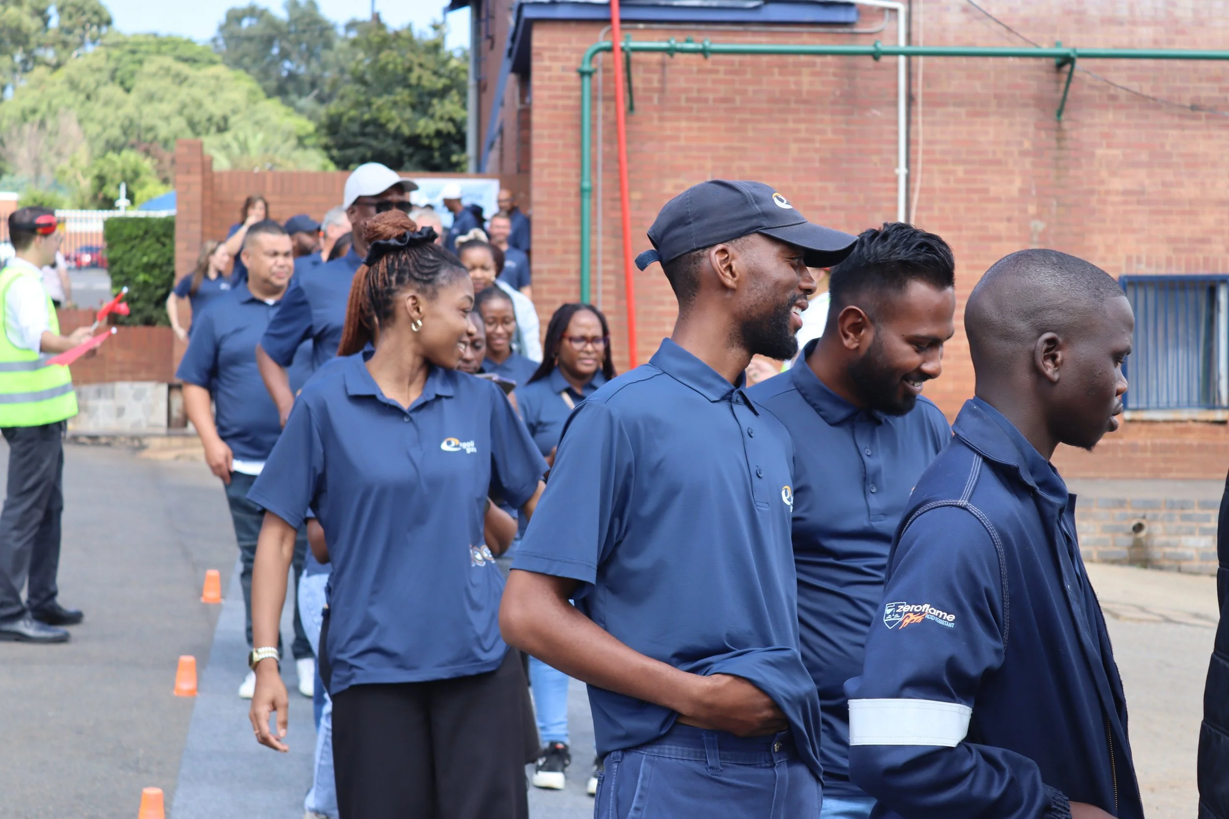 Group of people standing in a line outdoors during daytime, wearing blue shirts, some smiling, and cones on the ground.