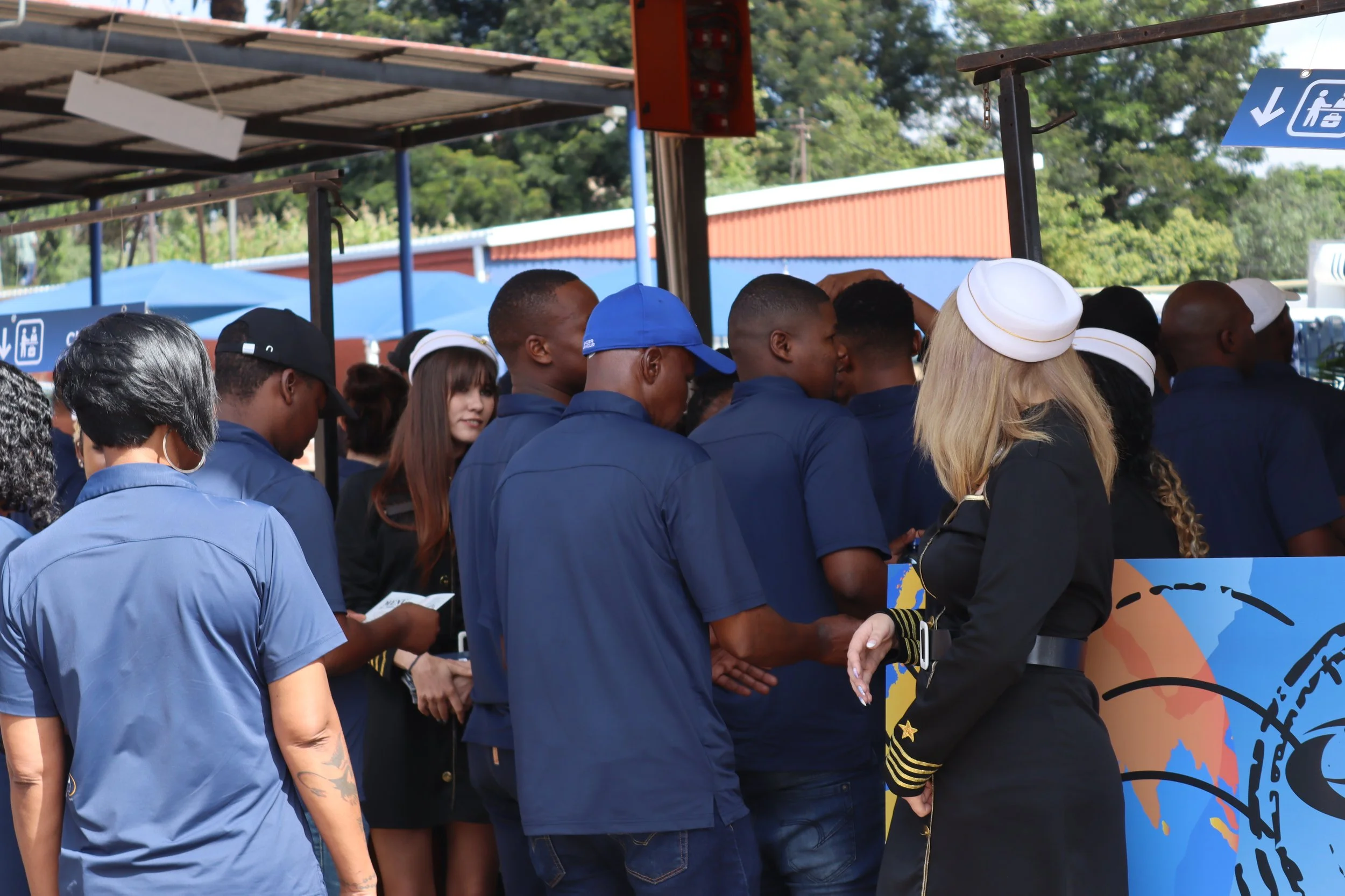 A group of people, many in blue uniforms and some wearing hats, are gathered in line or engaged in conversation outdoors under a shaded area. There are trees and a building with an orange wall in the background, along with blue umbrellas and signs wi