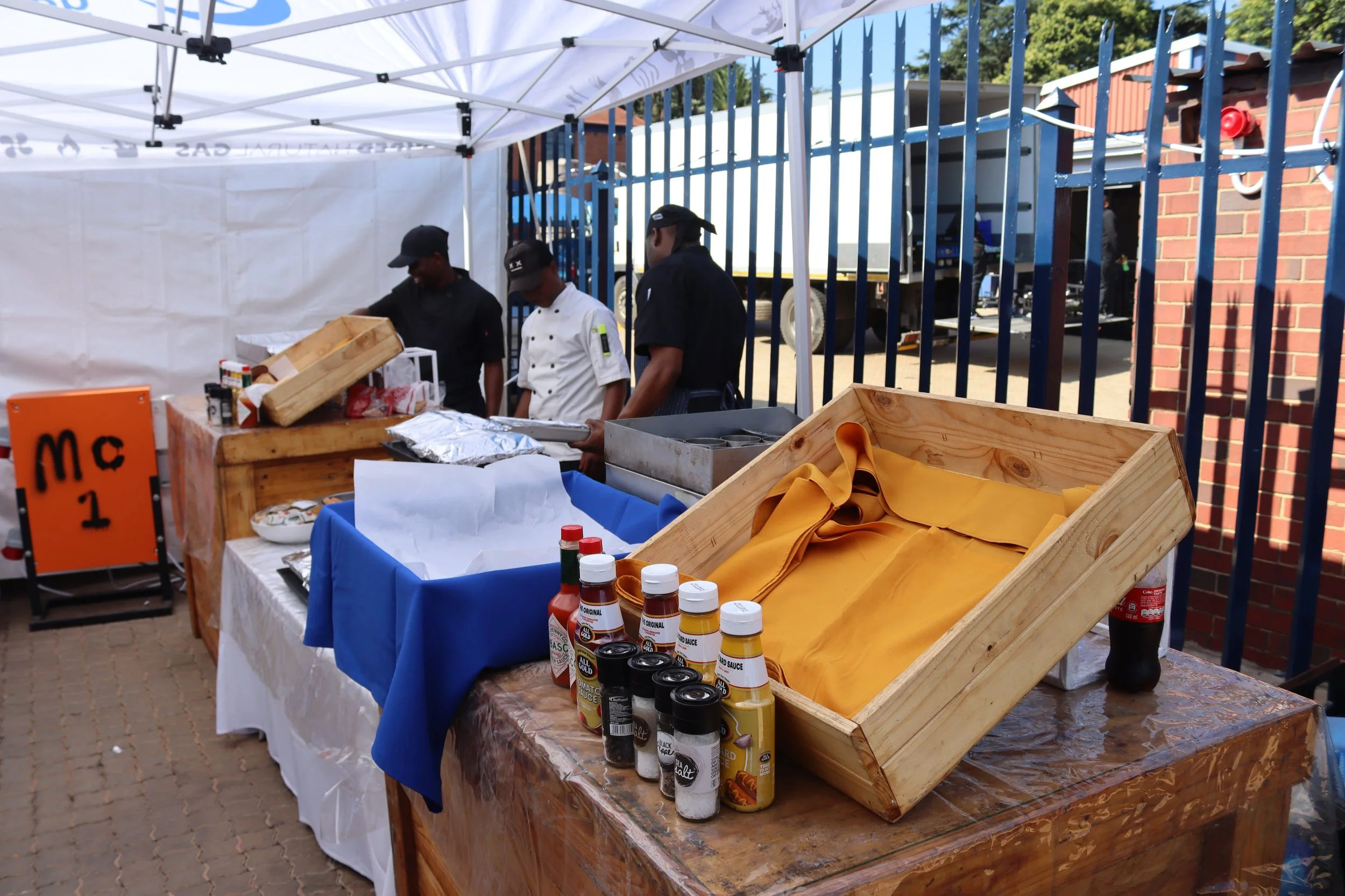 Food stall with condiments and supplies, staff preparing food, set up outdoors with a tent and a brick wall in the background.