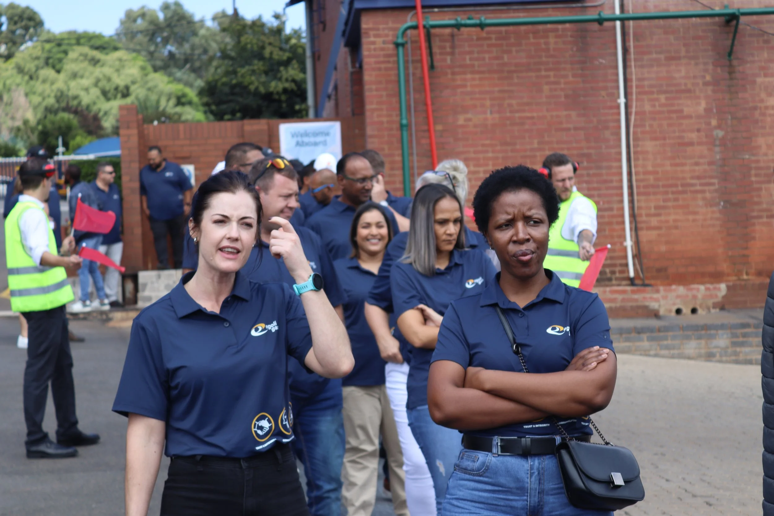 A group of people wearing navy blue shirts with logos gathered outdoors near a brick wall, some with serious expressions, some smiling, in a casual setting.