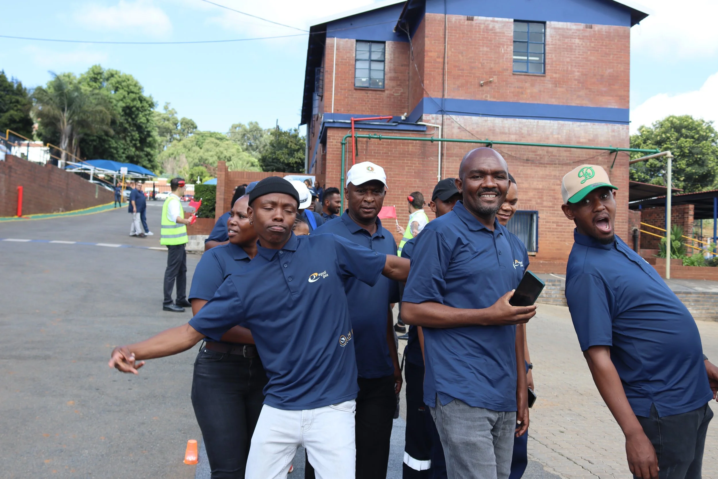 Group of people standing outside near a brick building, some holding hands, wearing dark blue uniforms, and facing the camera, with a few smiling.