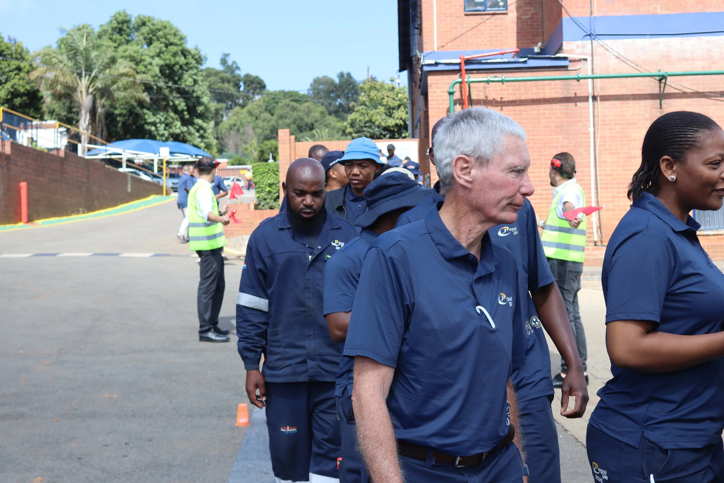 A group of people in navy blue uniforms walking in line outdoors near a brick building and lush green trees, with some officials wearing neon vests in the background.