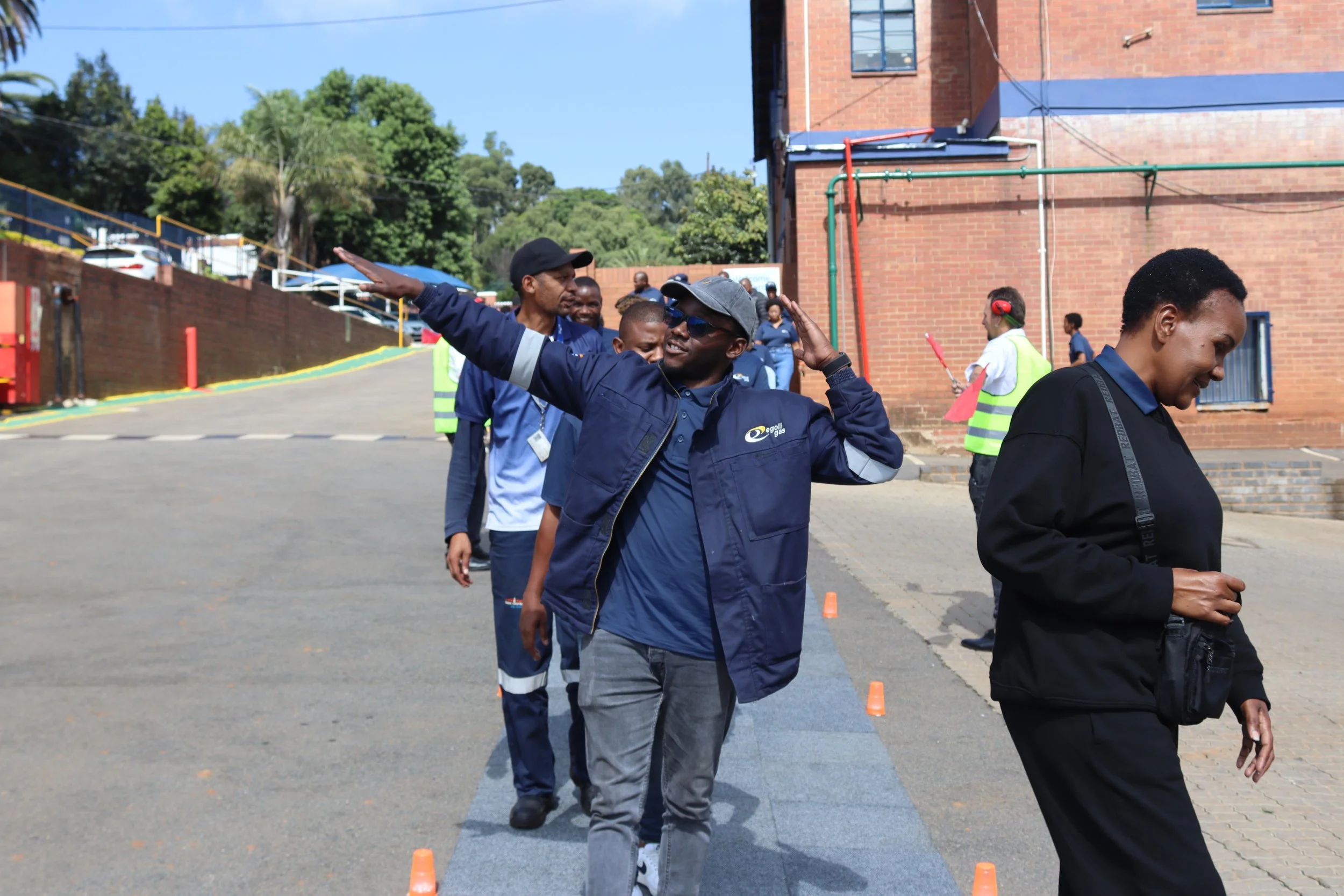 A group of people are outdoors, some wearing safety vests, participating in an event involving orange traffic cones on a sunny day. One person is doing a gesture with his hand, and others are smiling or looking down, with a brick building and trees i