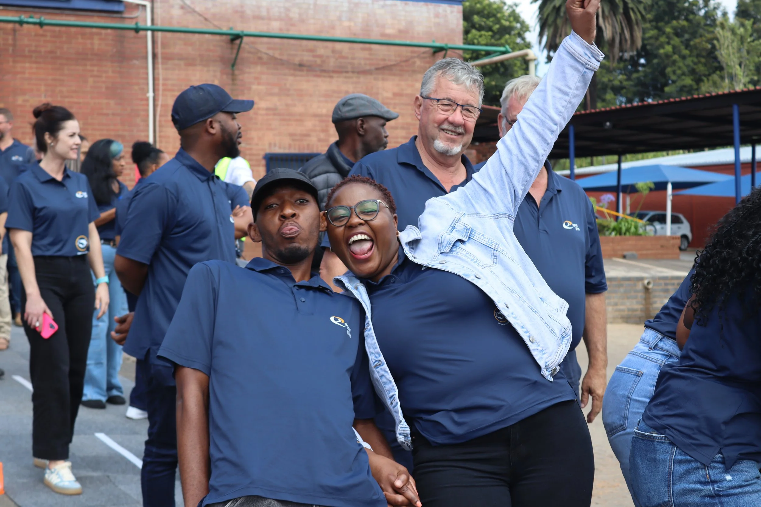 Group of diverse people outdoors, some wearing navy blue shirts with a logo, smiling and raising their hands in a celebratory or political event. Background includes a brick building and green trees.