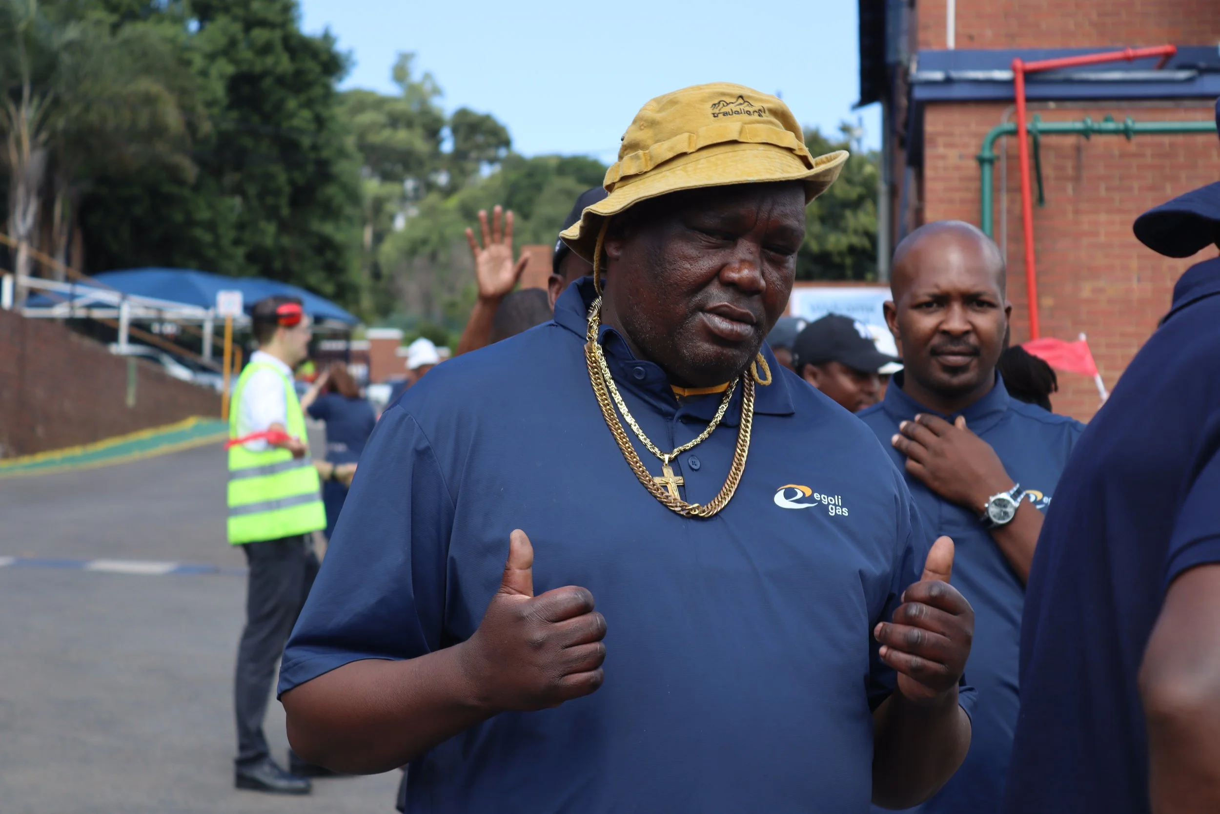A man in a yellow hat and blue shirt with the Egoli Gas logo, wearing gold chains and giving a thumbs-up, standing outdoors among other people.