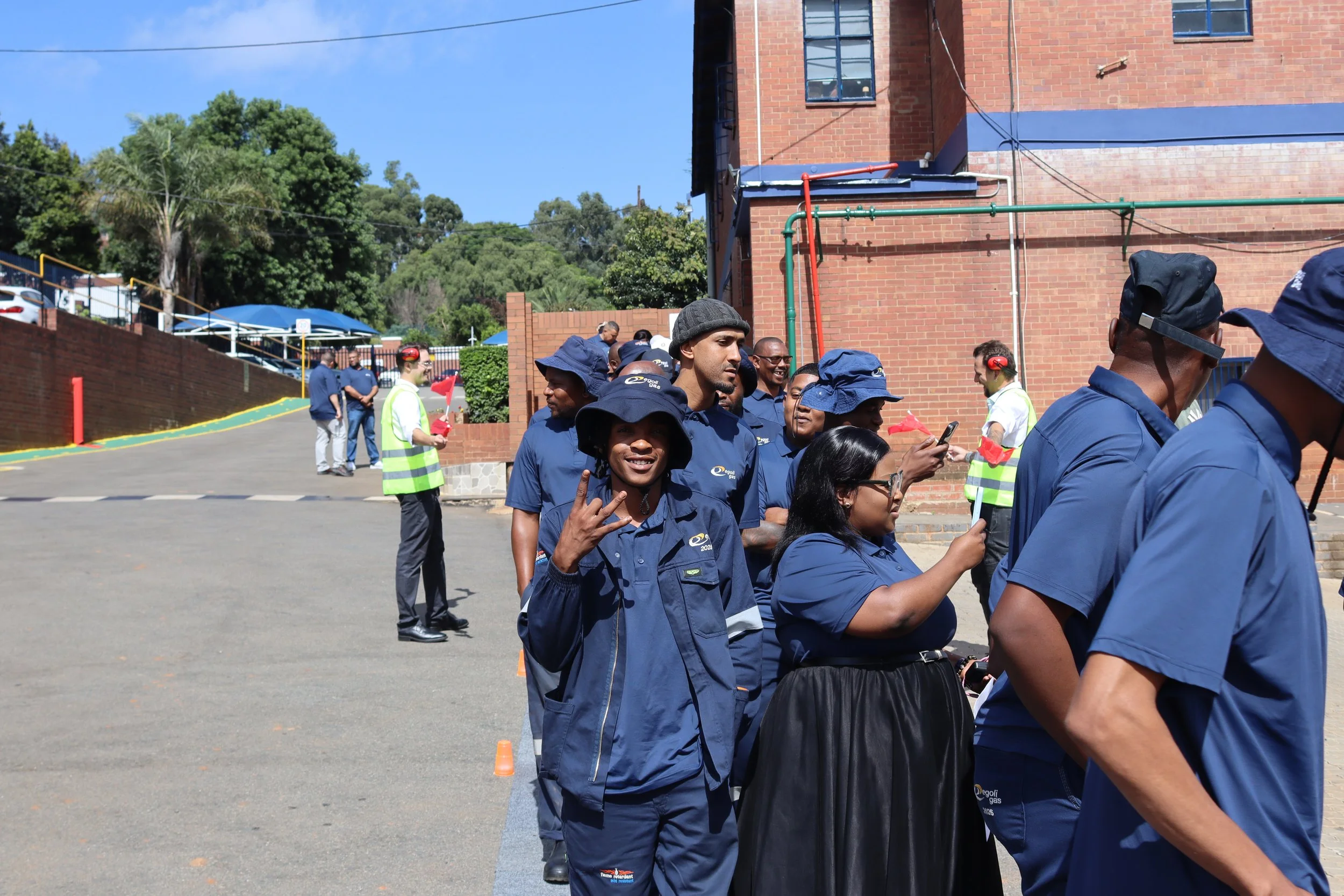 Group of people standing in line outdoors on a sunny day, some wearing blue uniforms with 'Egol Gas' logos, others in casual clothing, some with hats and headphones, near a brick building with green and red pipes, trees, and a blue sky.