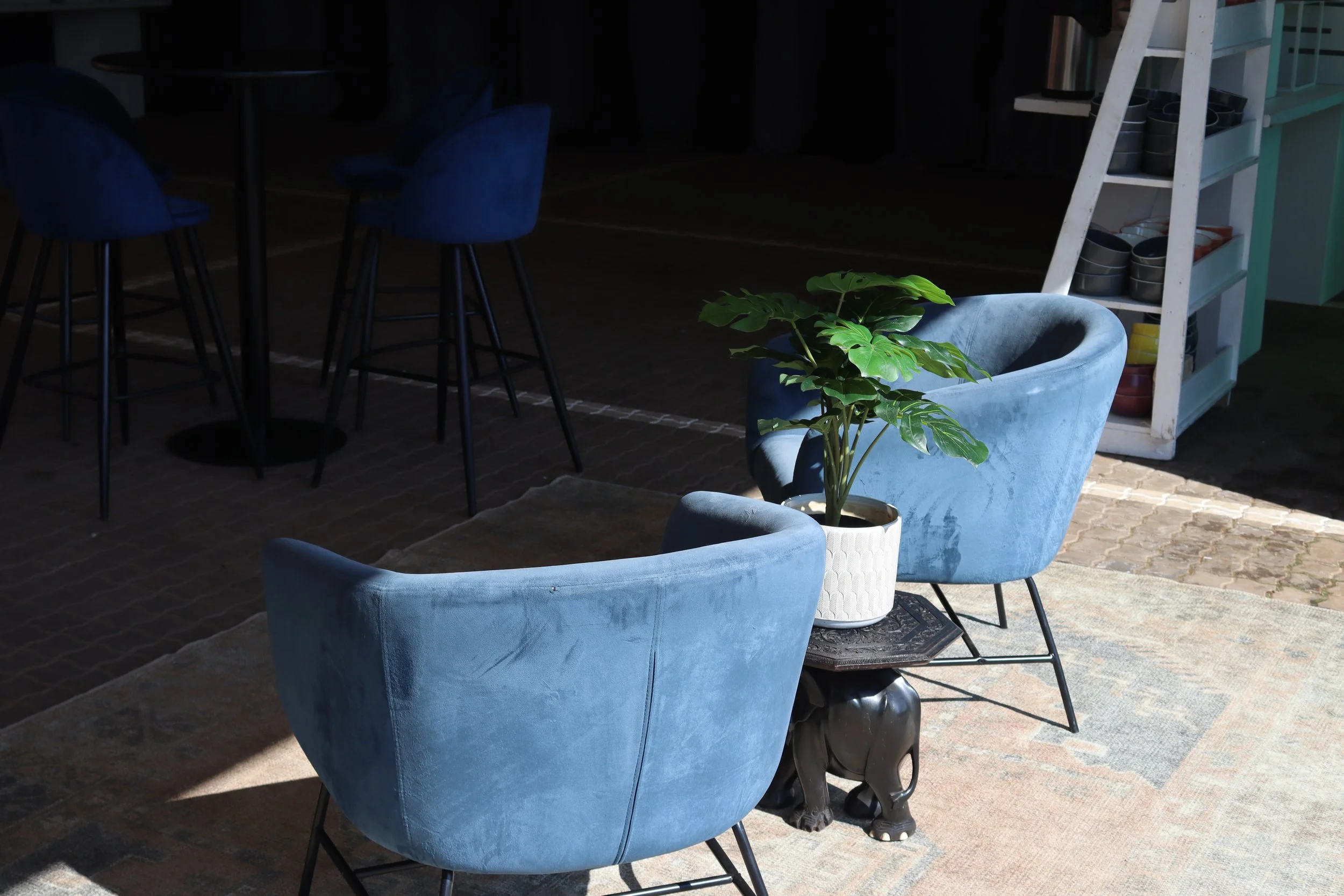 Outdoor seating area with blue velvet armchairs, a potted plant, a white shelving unit with black and white bowls, and tall blue bar stools, with sunlight casting shadows on the brick floor.