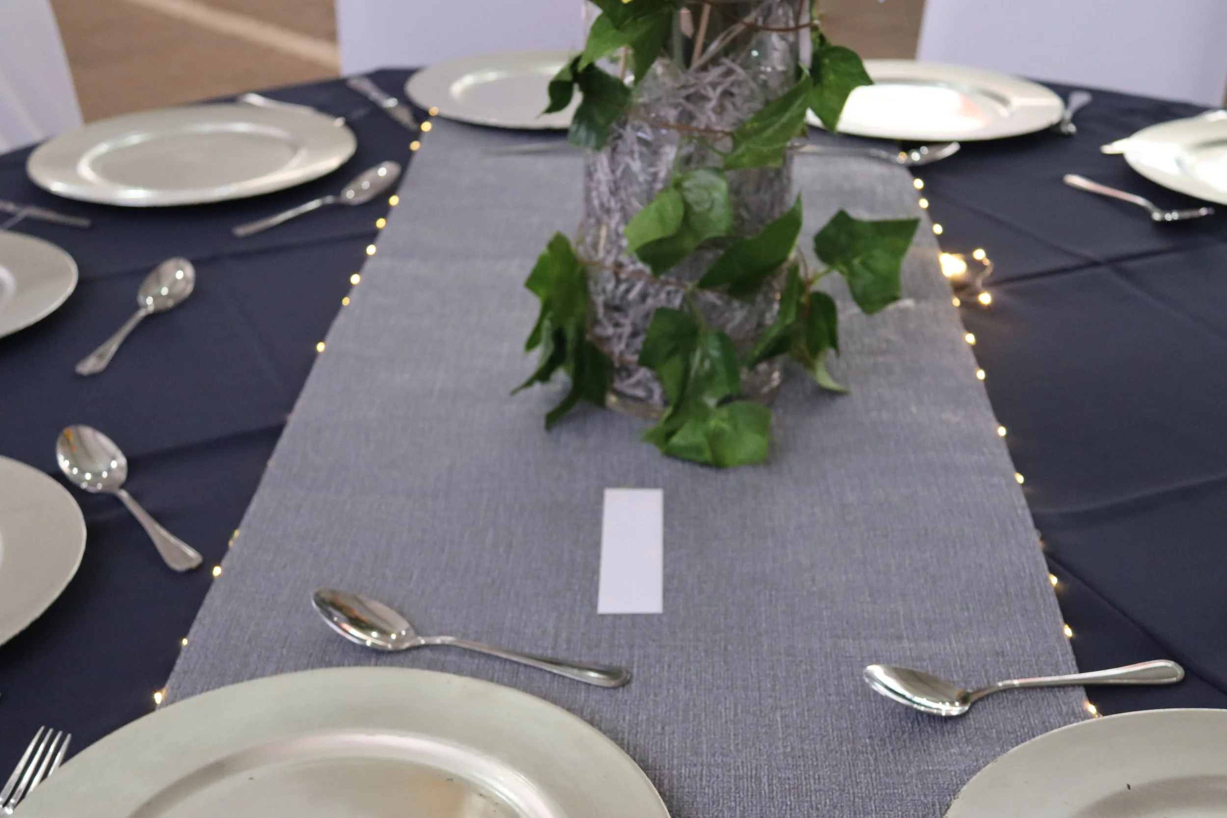 A formal dining table with a centerpiece of a glass vase filled with greenery, set with white plates, silverware, and a table runner, surrounded by chairs.