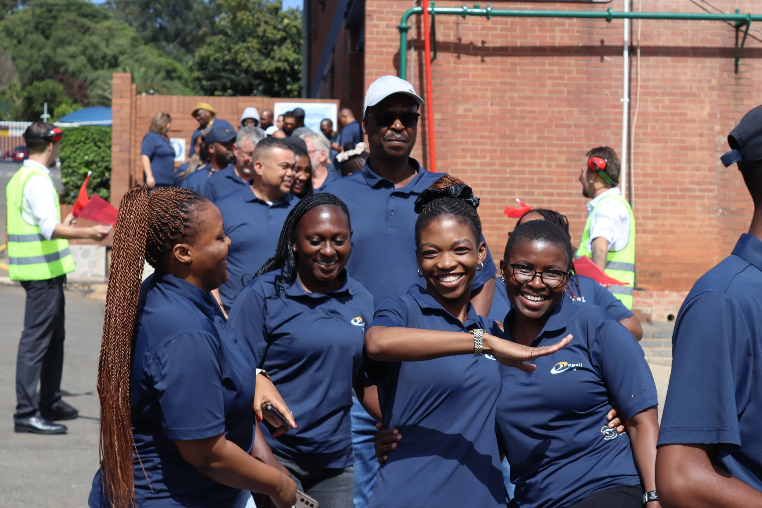 A group of people wearing matching navy blue shirts gathered outdoors, some smiling and engaging in a handshake or greeting. In the background, there are individuals with headsets and a brick building with green piping, under a sunny sky with trees.