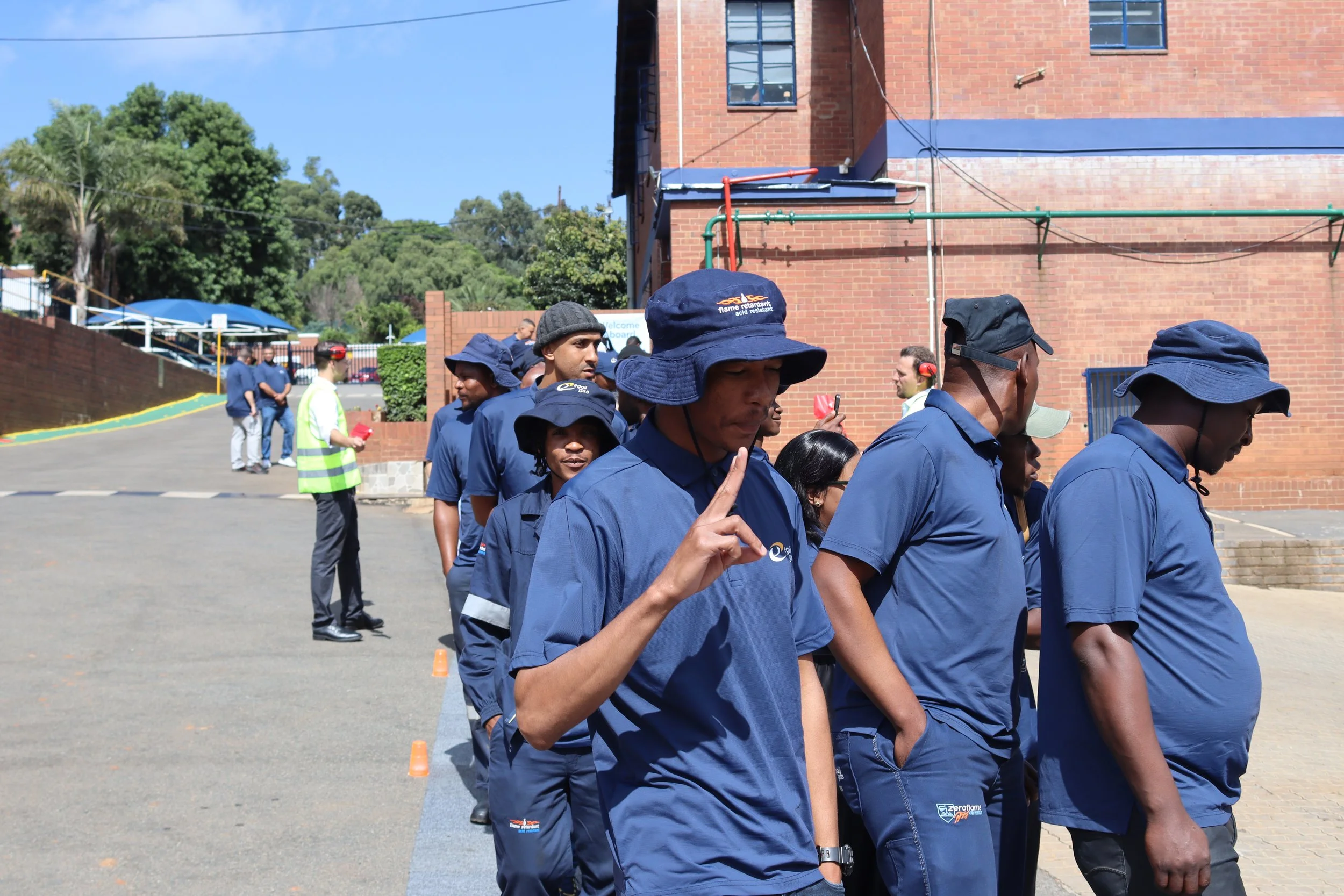 Line of people walking outdoors near a brick building, some wearing blue uniforms and hats, with a person in the foreground raising a hand.