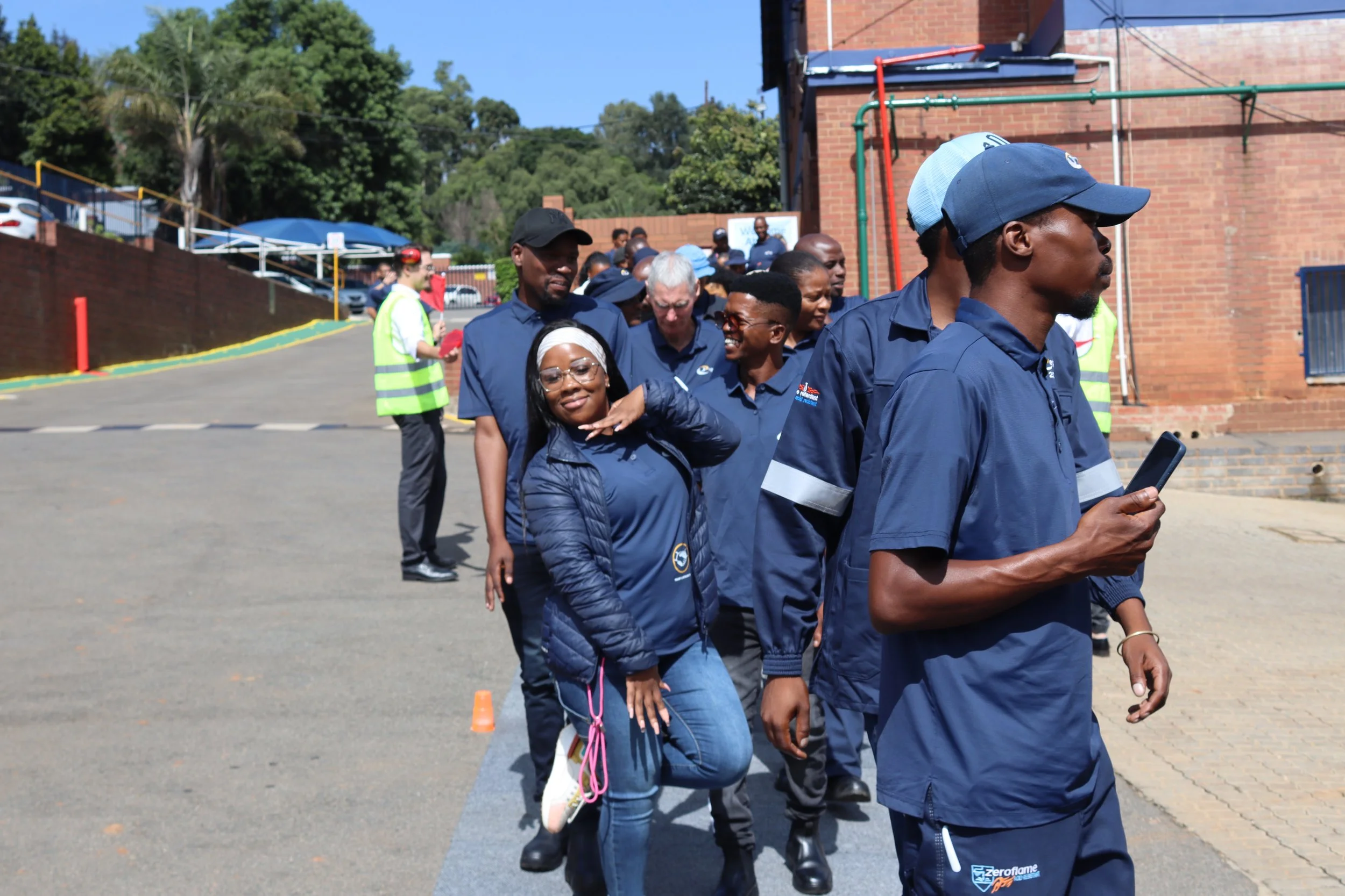 A group of people walking in a line outdoors, with some smiling and one person making a peace sign. They are dressed in casual and uniform-like clothing, and the background shows a brick building, trees, and a parking lot.