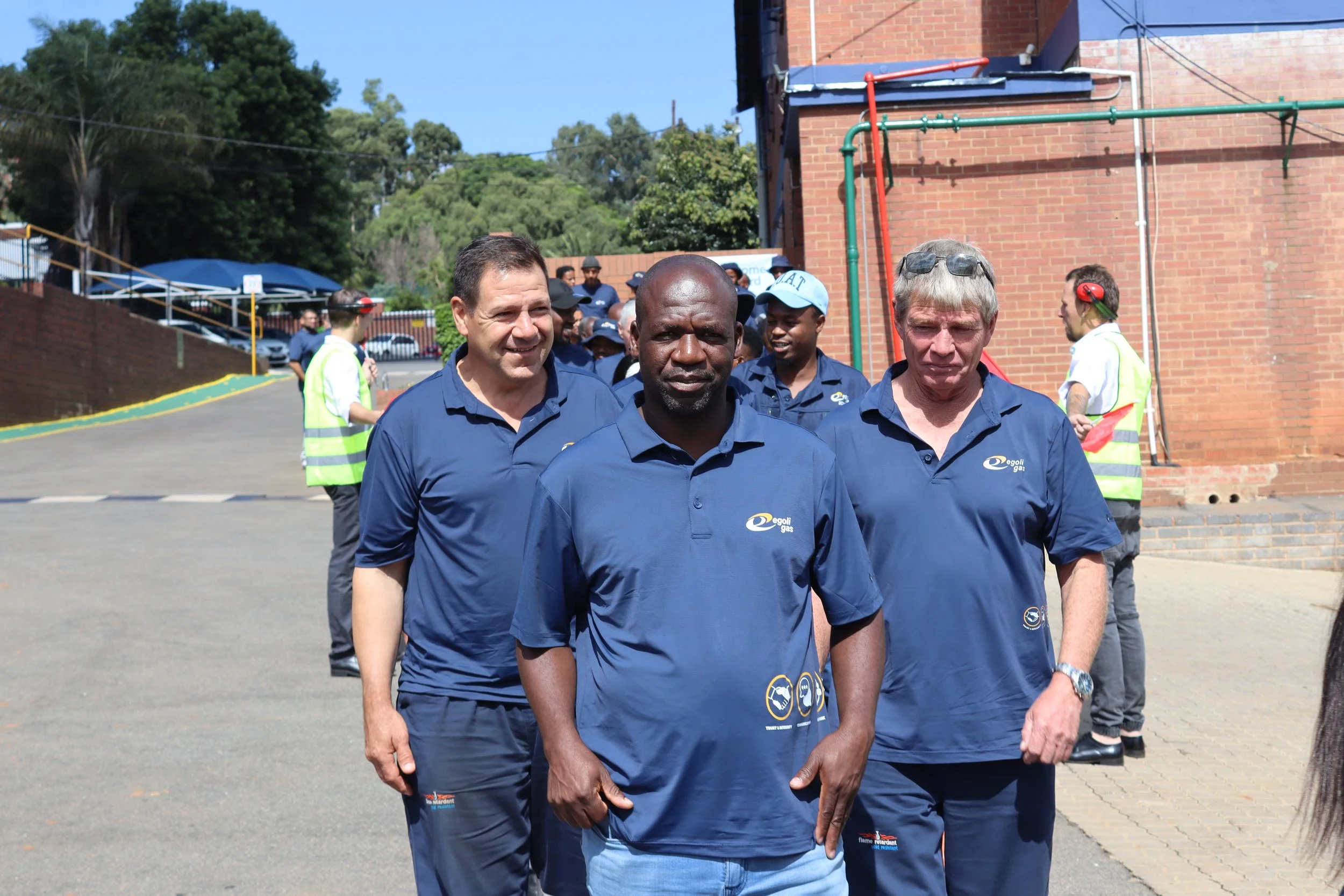 A group of diverse people walking outside together, wearing matching blue shirts with a logo, with a brick building and trees in the background.