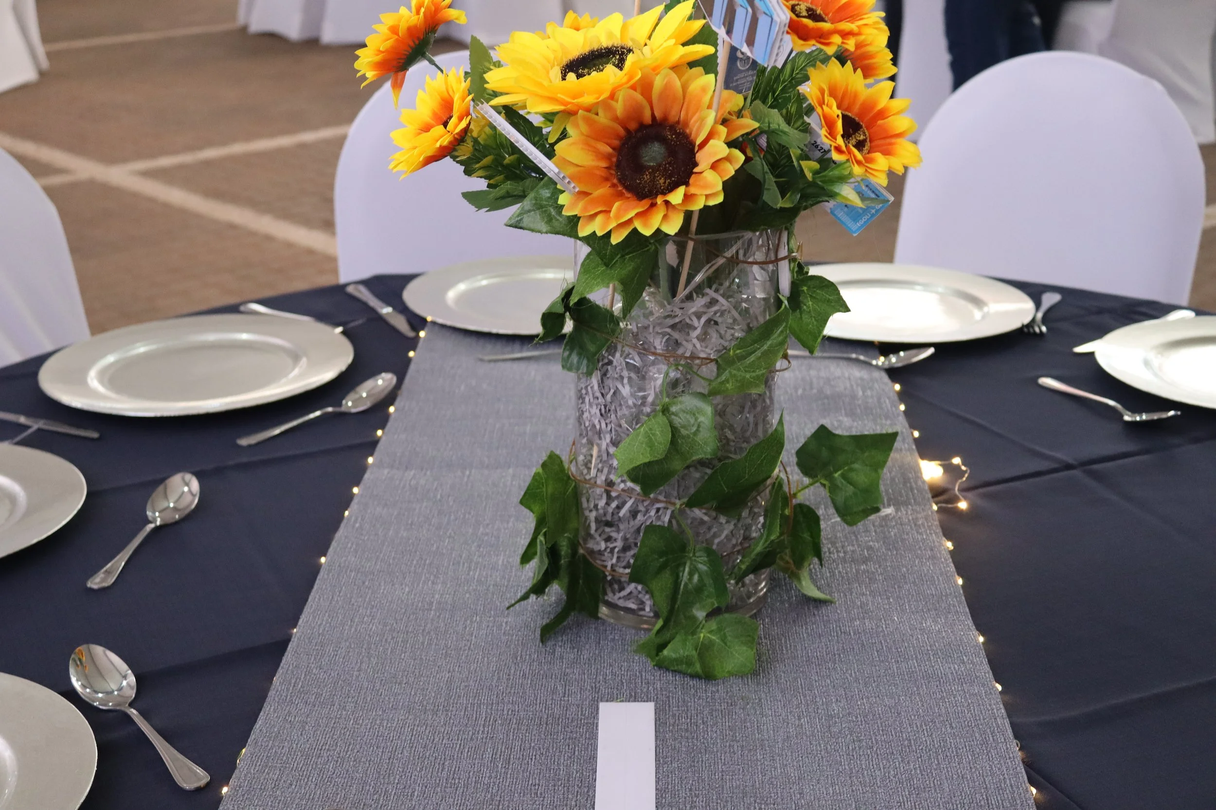 A table setting with a centerpiece of yellow and orange sunflowers in a glass vase with green leaves, surrounded by white plates, silverware, and a gray table runner, with chairs and a blurred background.