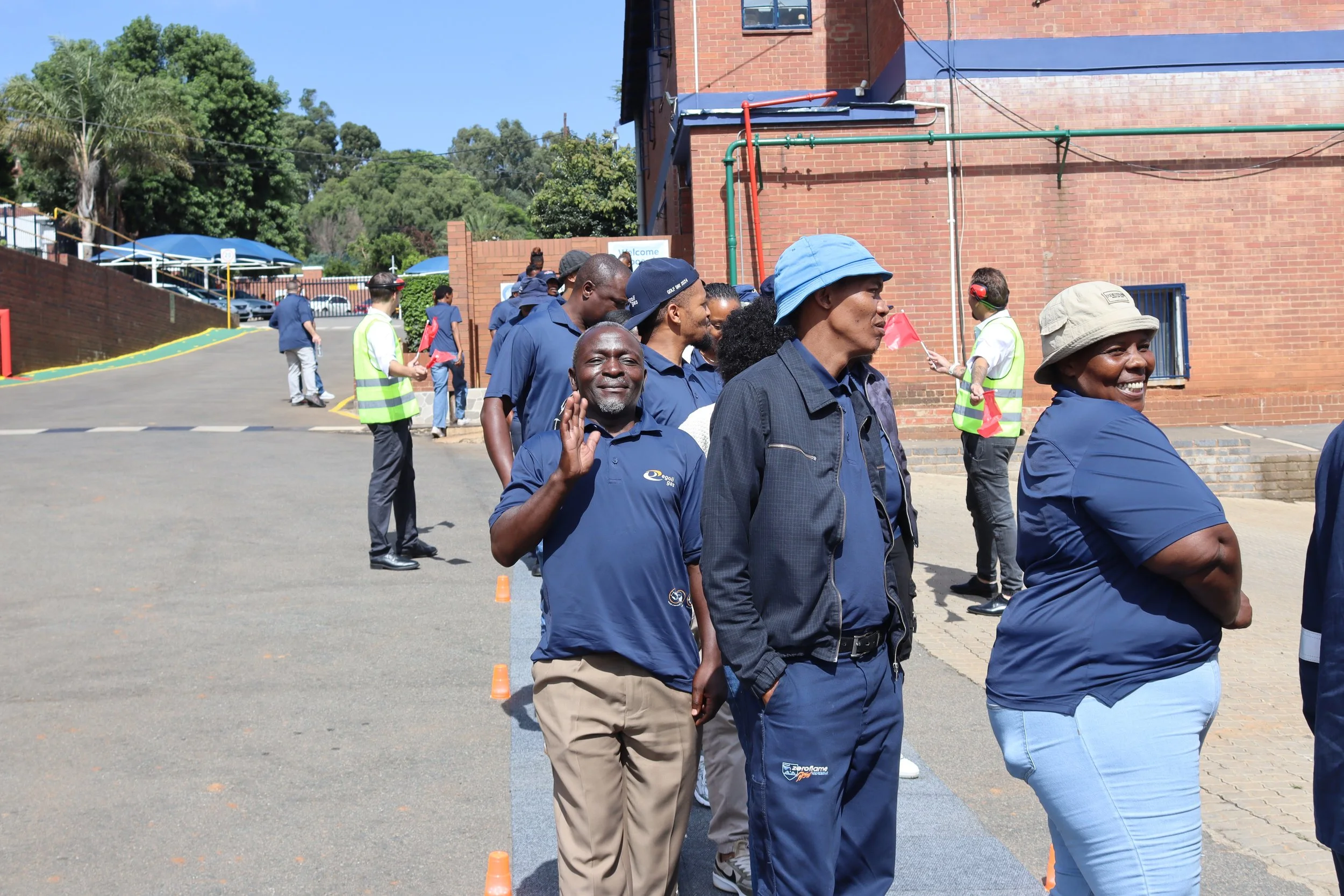 A group of diverse people standing in line outdoors on a sunny day, some wearing hats and casual clothing, next to a brick building with green piping and trees in the background.