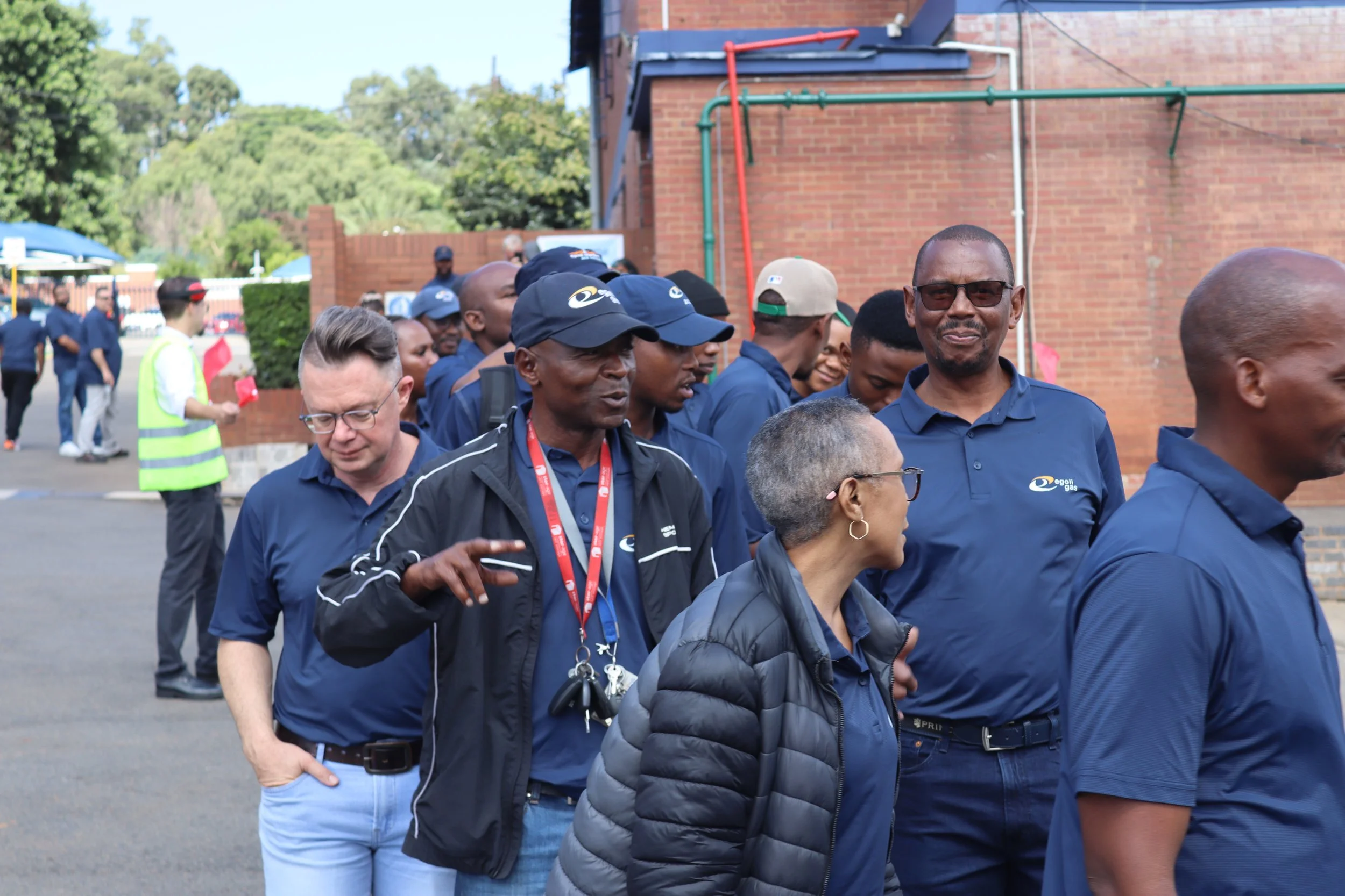 A group of people walking outdoors near a brick building, some wearing dark blue clothing and hats, with greenery and blue sky in the background.