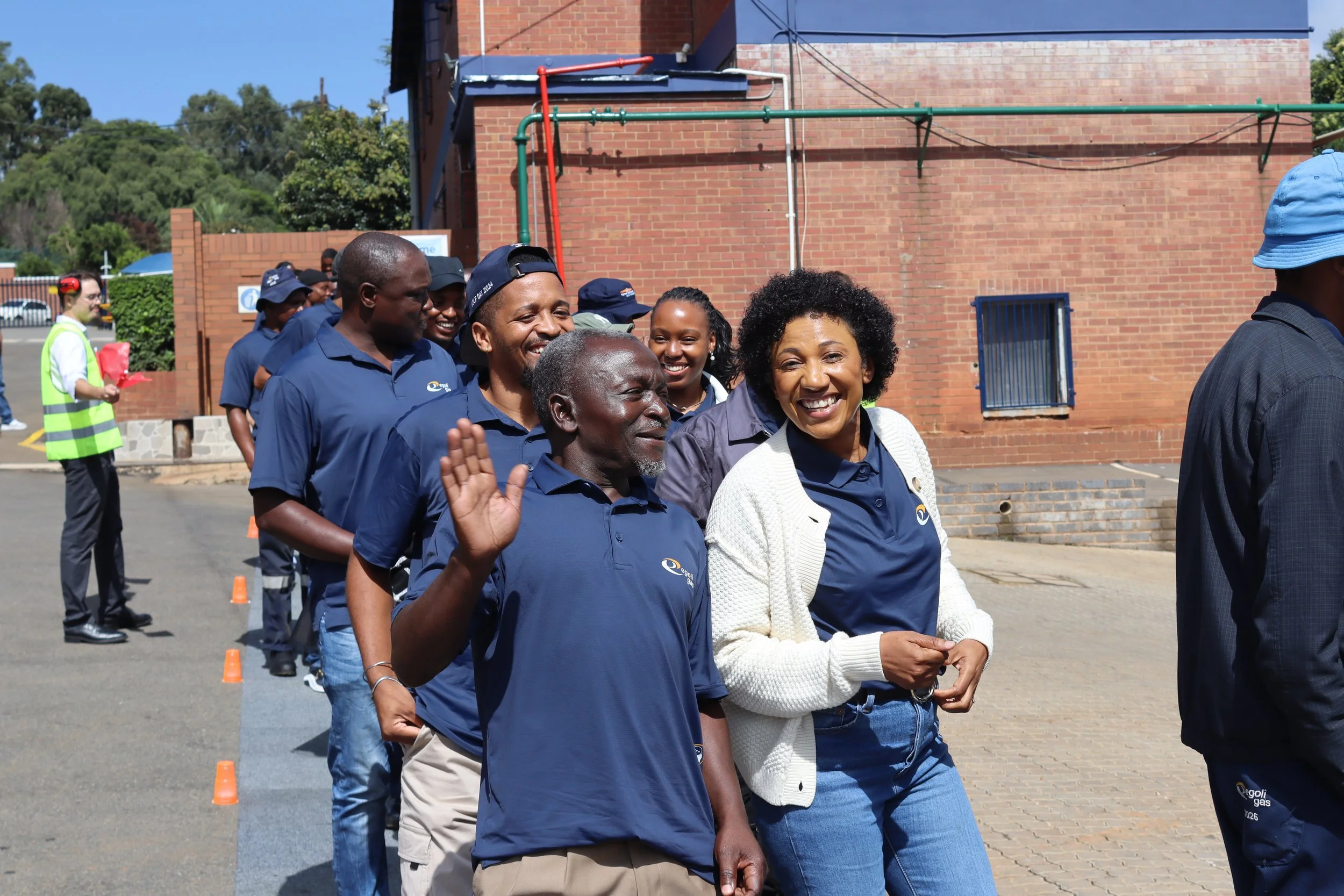 A group of diverse people smiling and waving while walking outdoors in front of a brick building, with some wearing blue shirts and hats, during a sunny day.