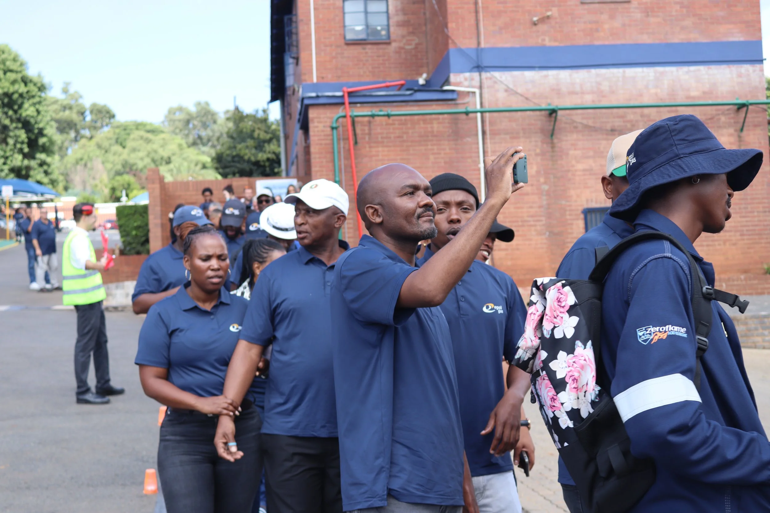 A group of people, mostly wearing blue shirts and some with hats, are standing outdoors near a brick building. Some are taking photos or videos, and a few are looking around or talking. Orange cones are placed on the ground, and trees are visible in 
