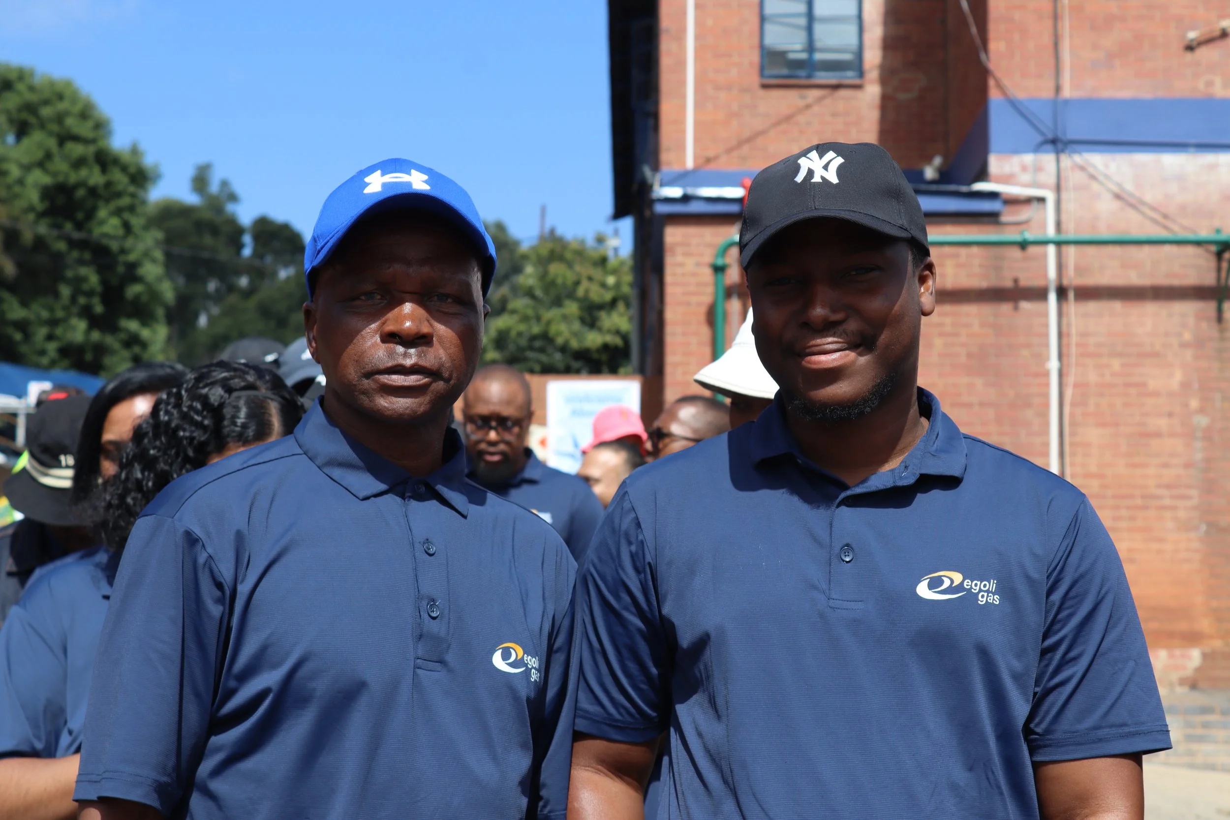 Two men wearing blue and black caps with the Egoli Gas logo, standing outdoors in front of a brick building and other people, possibly at an event or gathering.