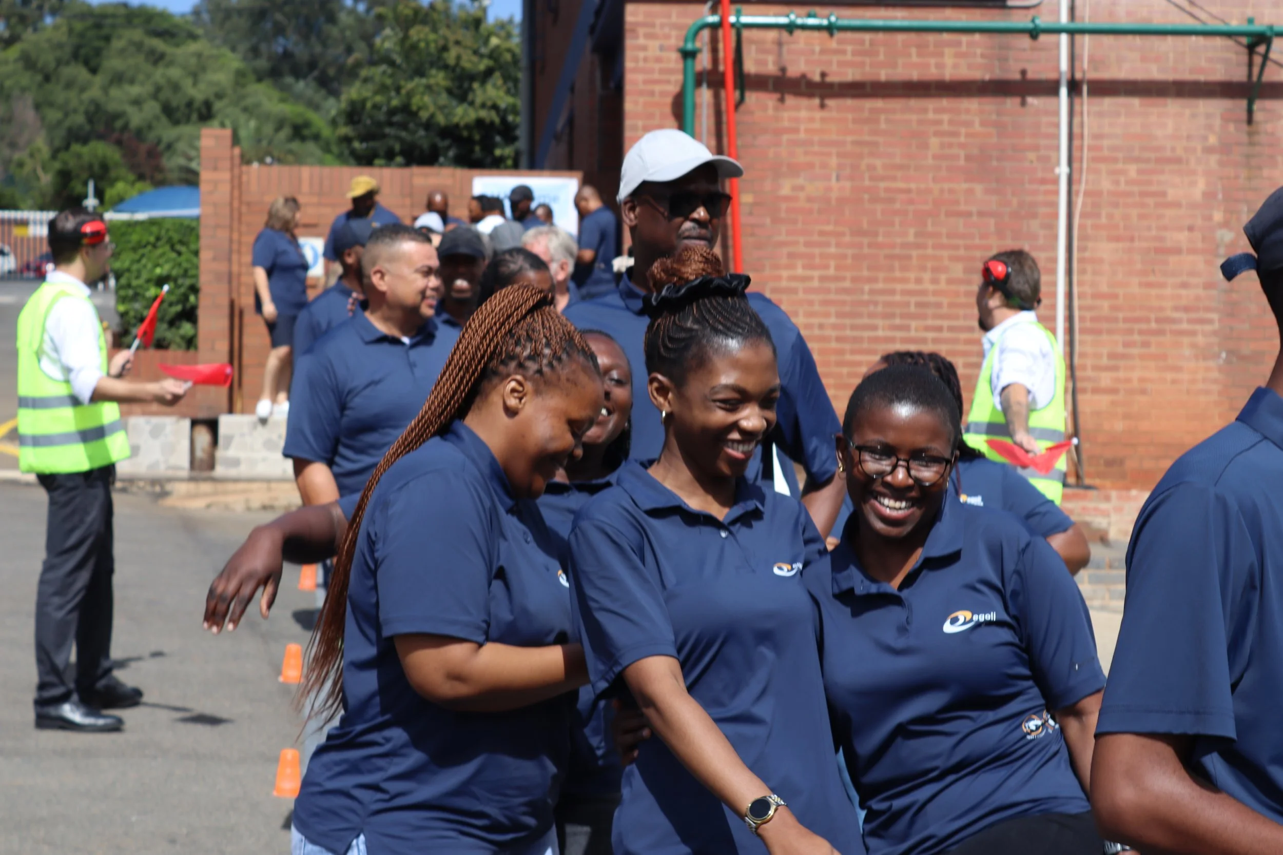 Group of smiling people in blue shirts participating in an outdoor team-building activity, with neon cones set up on the ground, brick building, and trees in the background.